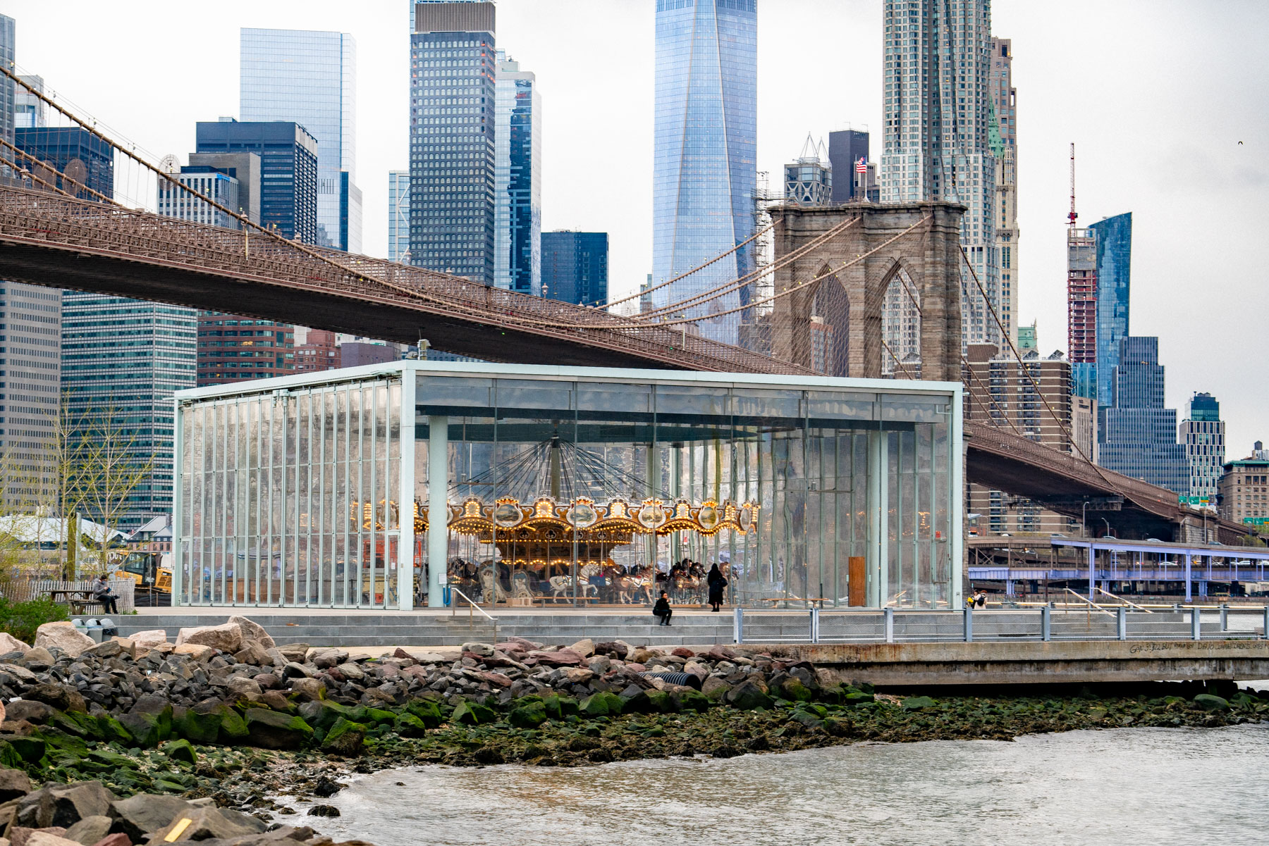 Jane's Carousel sits in its glass pavilion beneath the Brooklyn Bridge with the Manhattan skyline behind it.