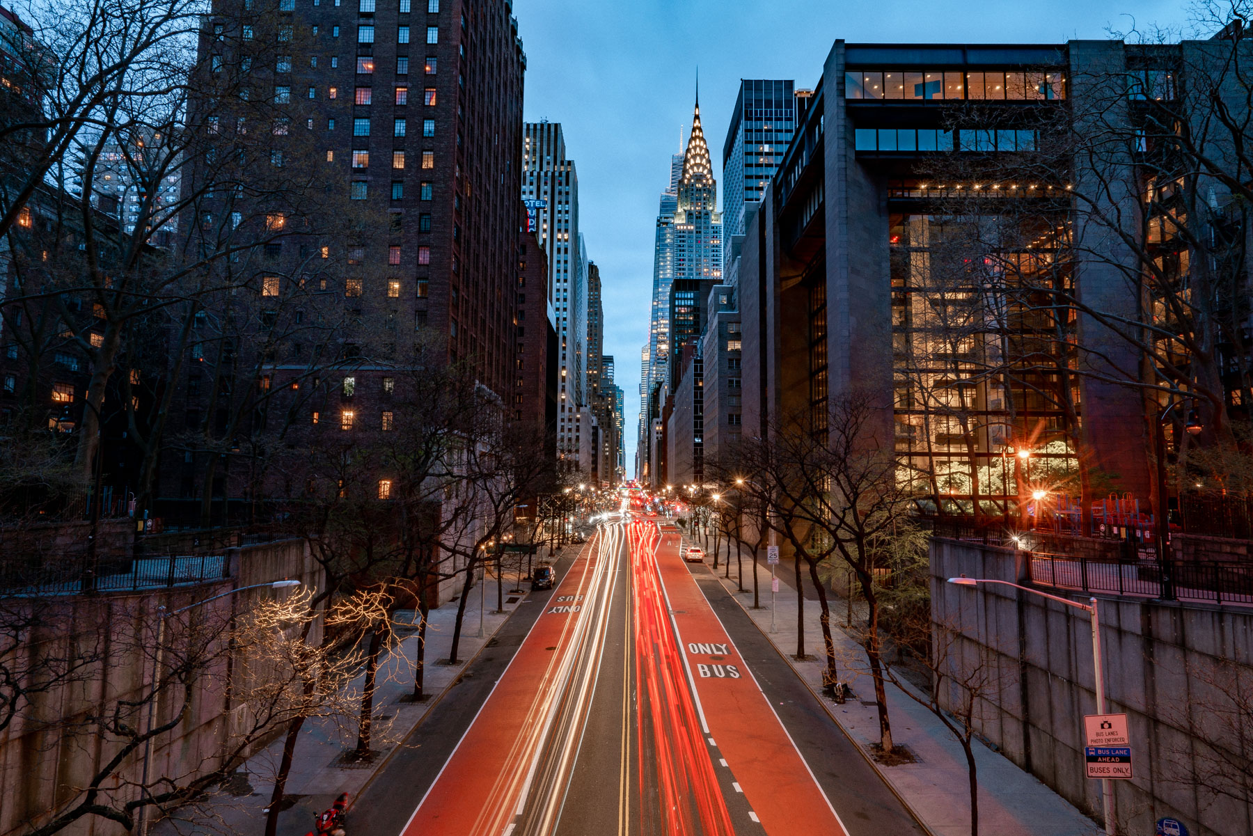 Long-exposure traffic light trails stream down 42nd Street toward the Chrysler Building at dusk.