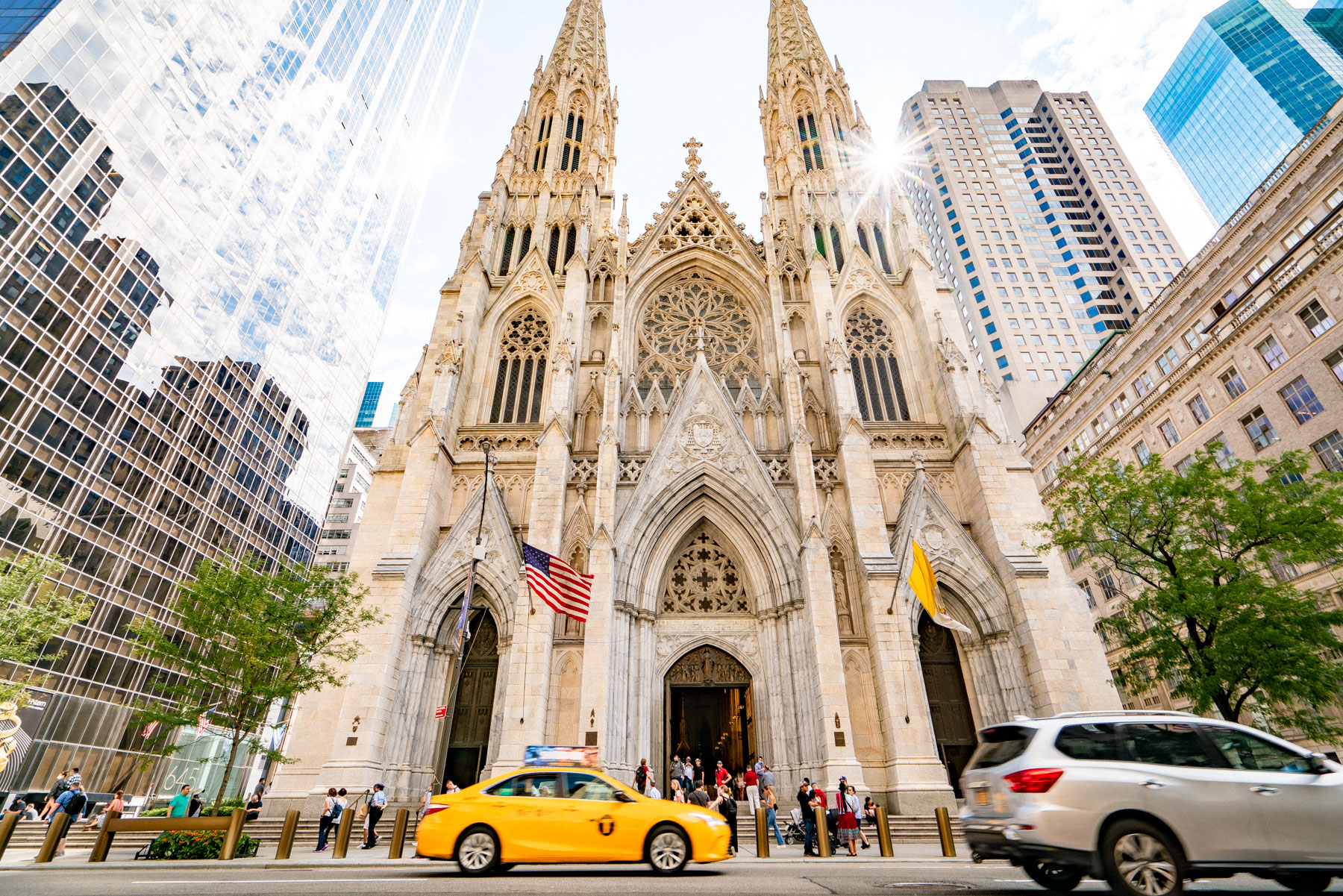 St. Patrick's Cathedral facade rises between skyscrapers on Fifth Avenue as a yellow taxi passes.