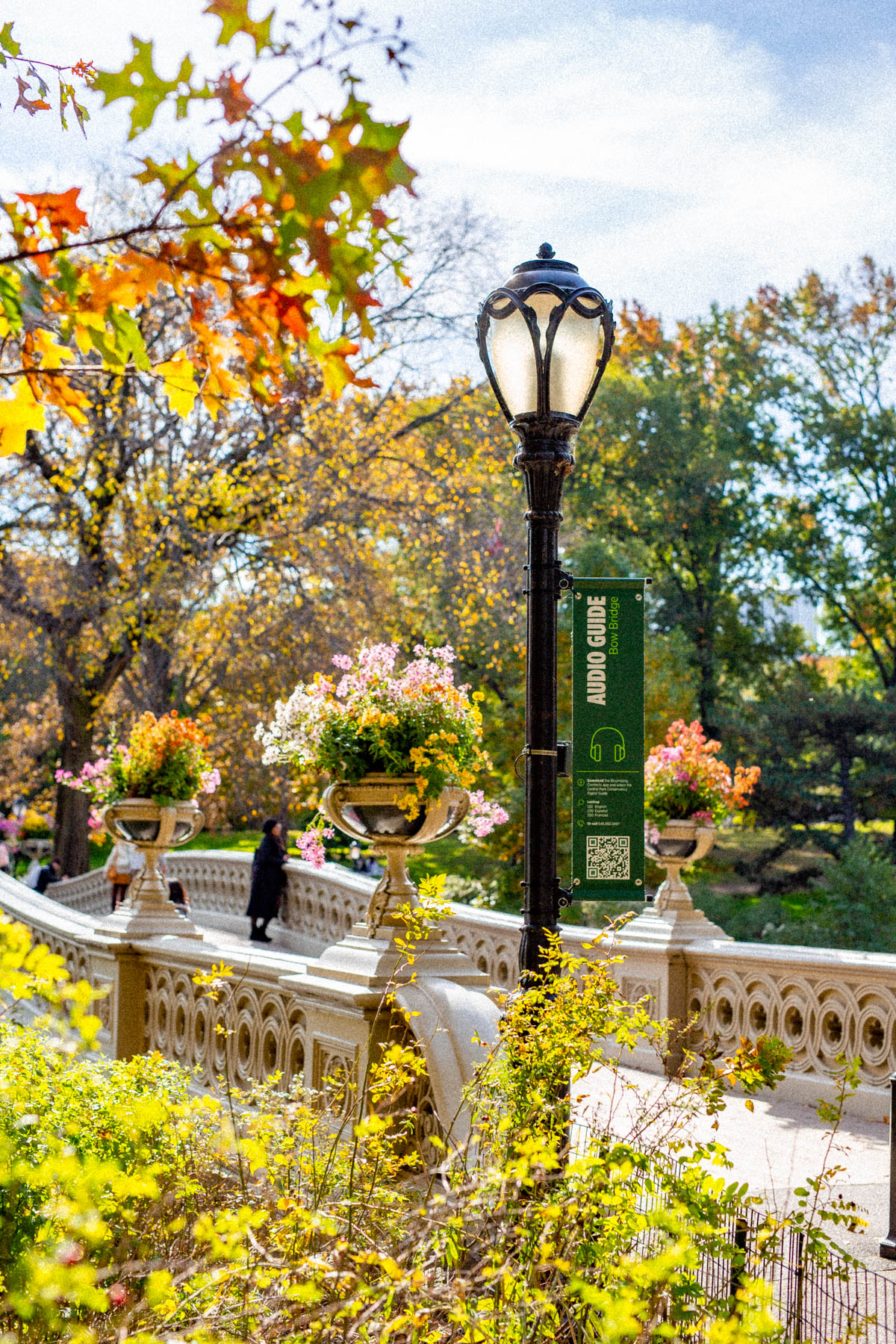 A lamppost with an audio guide sign stands on Bow Bridge in Central Park surrounded by fall foliage.