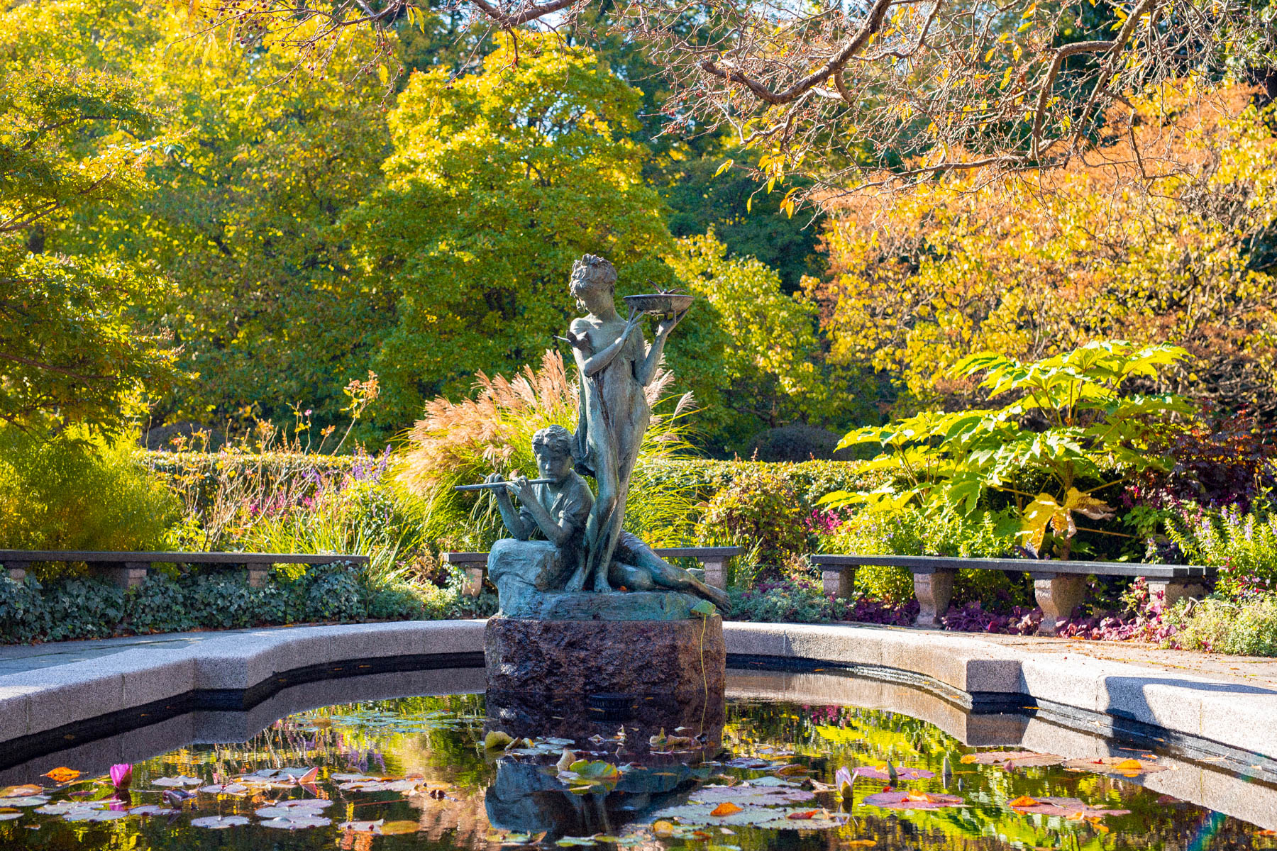 A bronze fountain statue stands amid a lily pond surrounded by autumn foliage in a park garden.