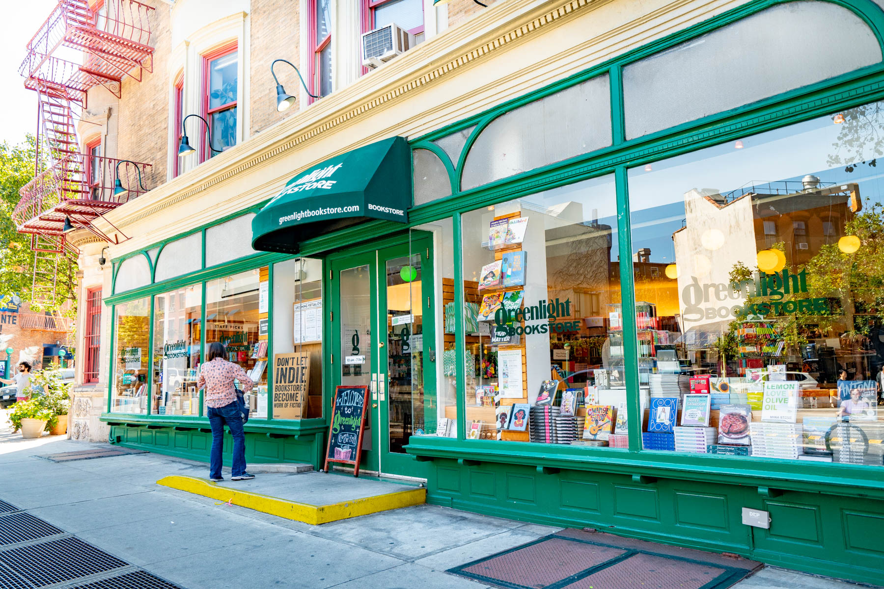 A green-awninged storefront of Greenlight Bookstore with a woman reading outside on the sidewalk.