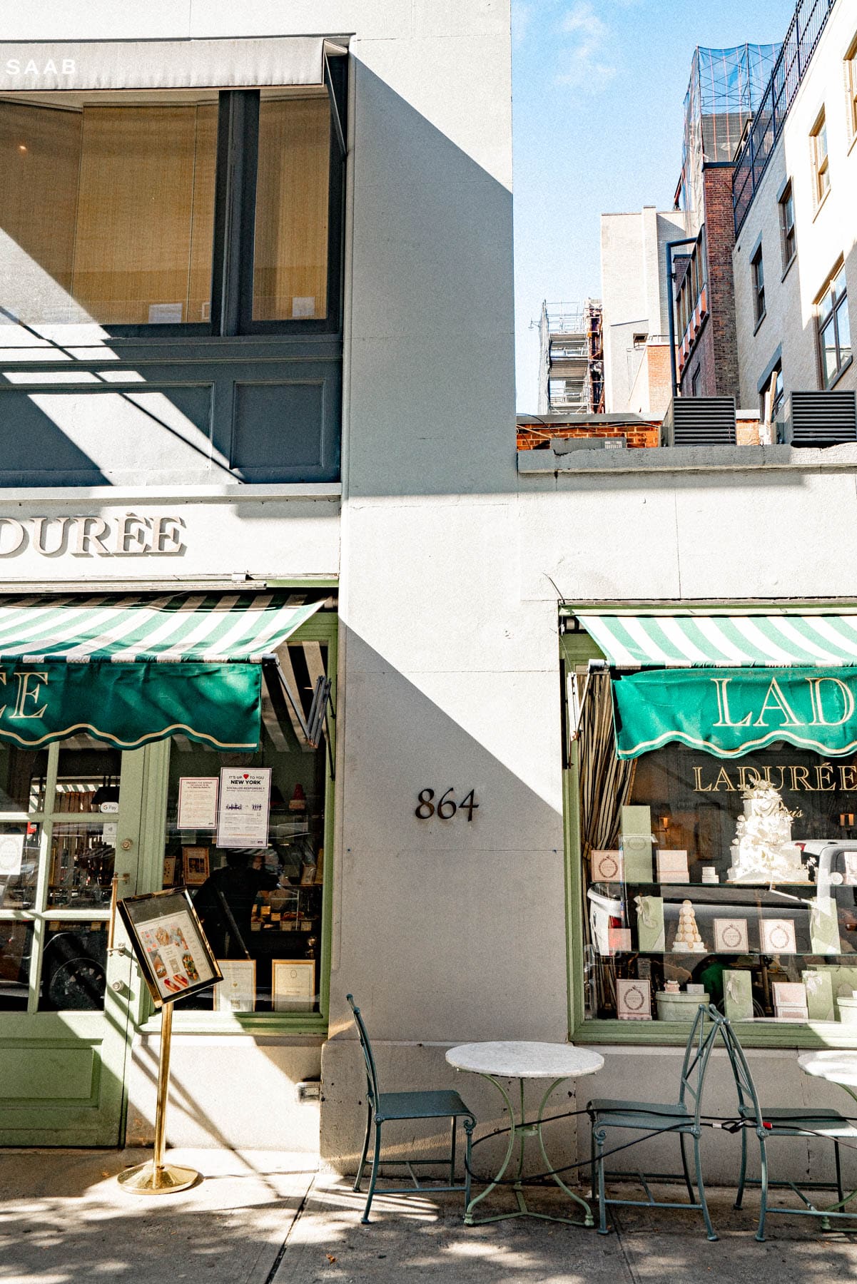 Storefront of Ladurée with green striped awnings and a sidewalk table on a sunny street corner.