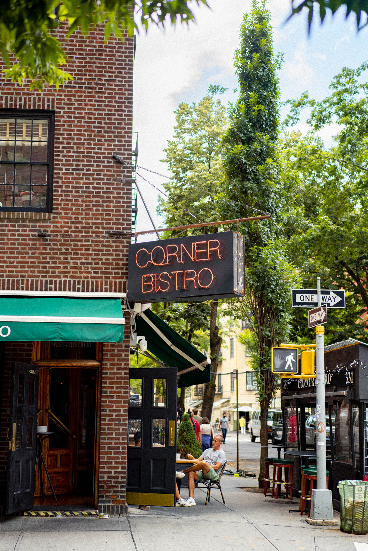 A neon Corner Bistro sign hangs above a West Village street corner with trees and a seated pedestrian.