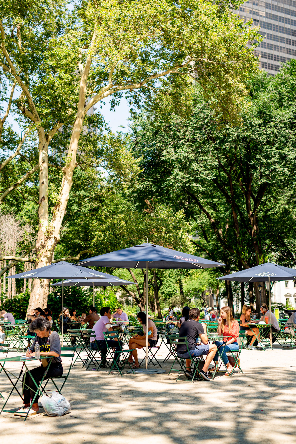 People sit at green tables under umbrellas in a shaded city park.