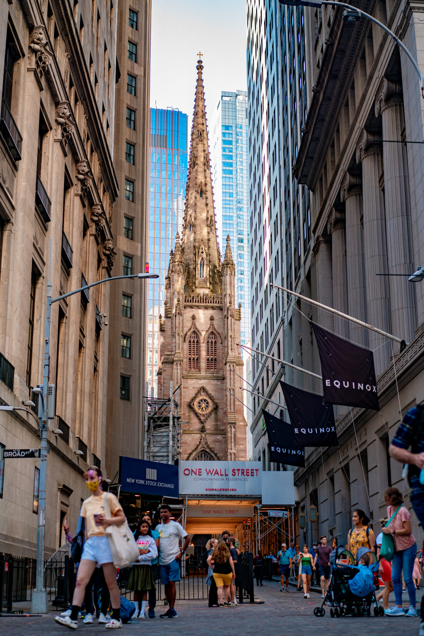 A view down Wall Street toward Trinity Church's spire, flanked by skyscrapers and pedestrians.