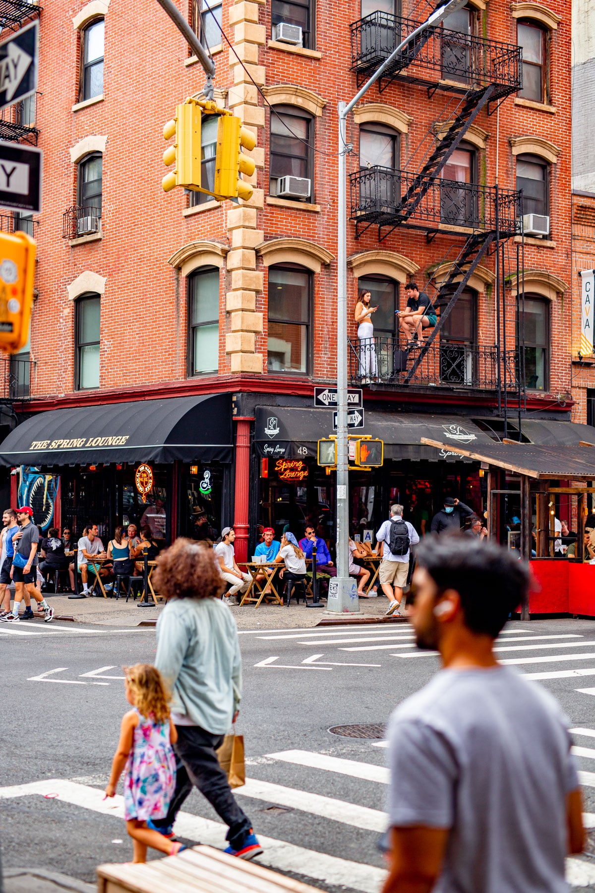 Pedestrians cross a Soho street corner beneath a brick building with a fire escape and the Spring Lounge bar below.