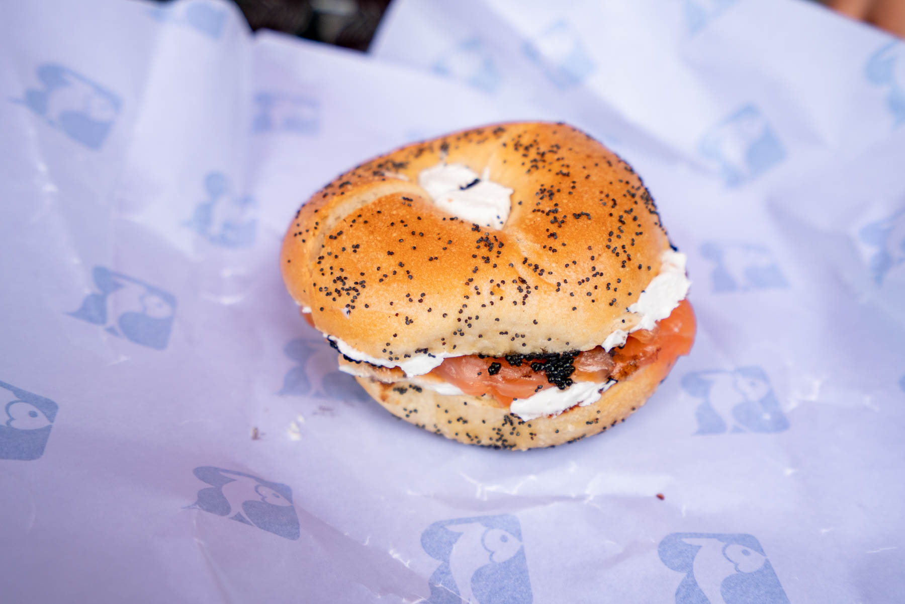 A poppy seed bagel with cream cheese and lox sits on wrapping paper printed with a fish logo.