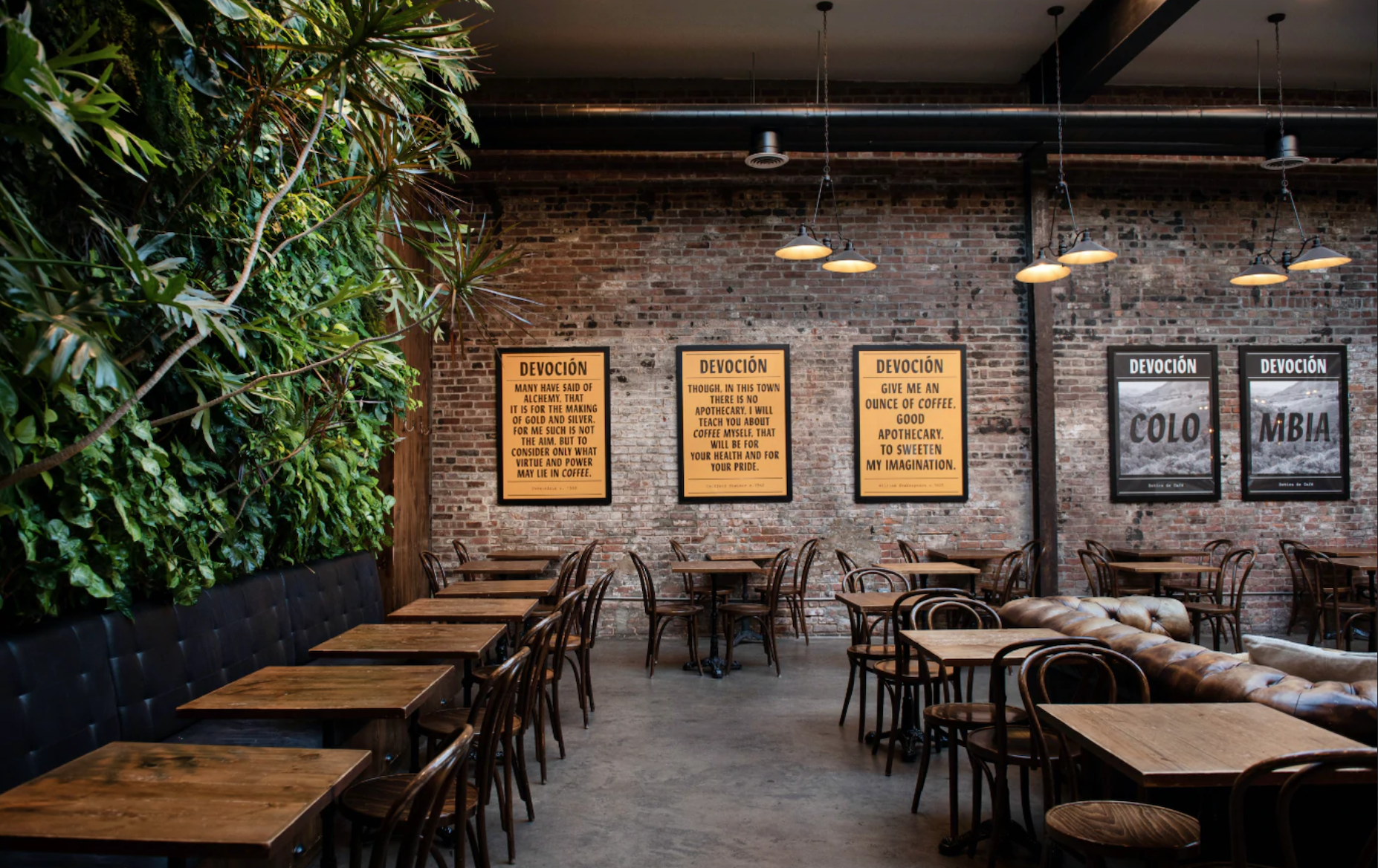 Interior of a coffee shop with exposed brick walls, framed posters, and a living plant wall alongside wooden tables and chairs.