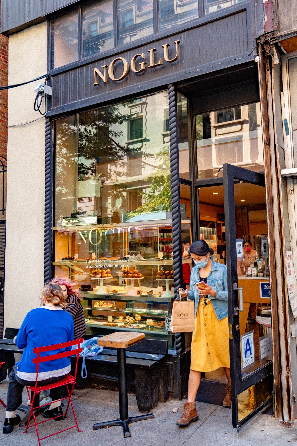 People sit and stand outside the gluten-free bakery NoGlu on a city sidewalk.