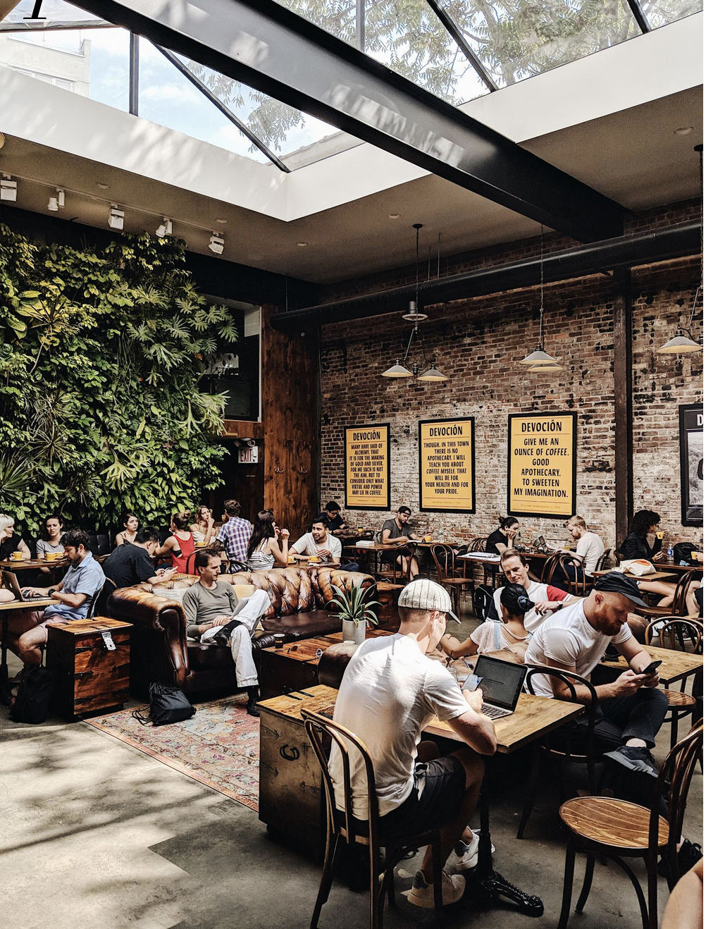 People sit and work at tables inside a plant-filled coffee shop with a skylight.