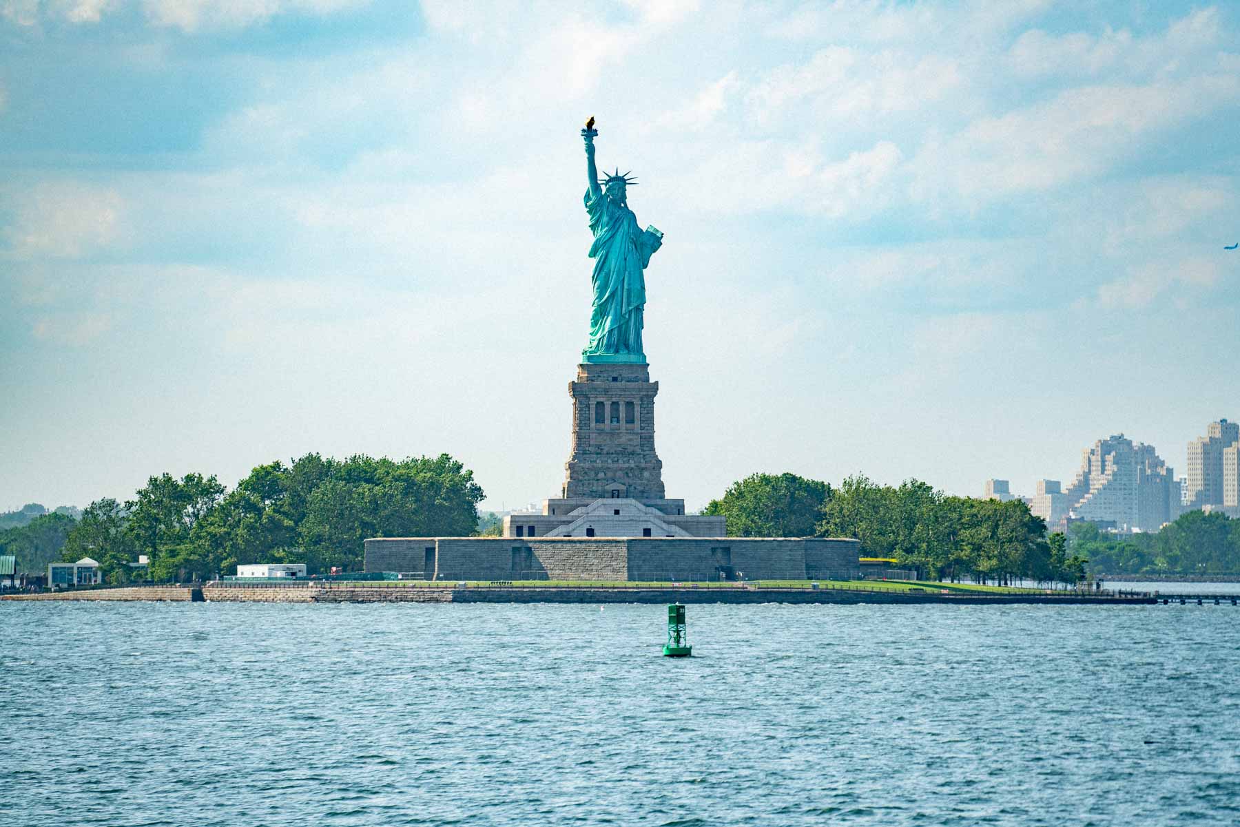 The Statue of Liberty stands on Liberty Island viewed from the water on a sunny day.