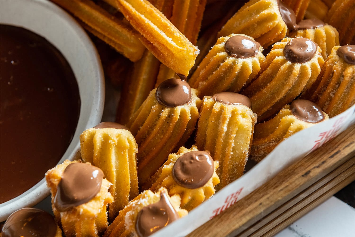 A basket of churros with chocolate dots sits beside a bowl of dulce de leche dipping sauce.