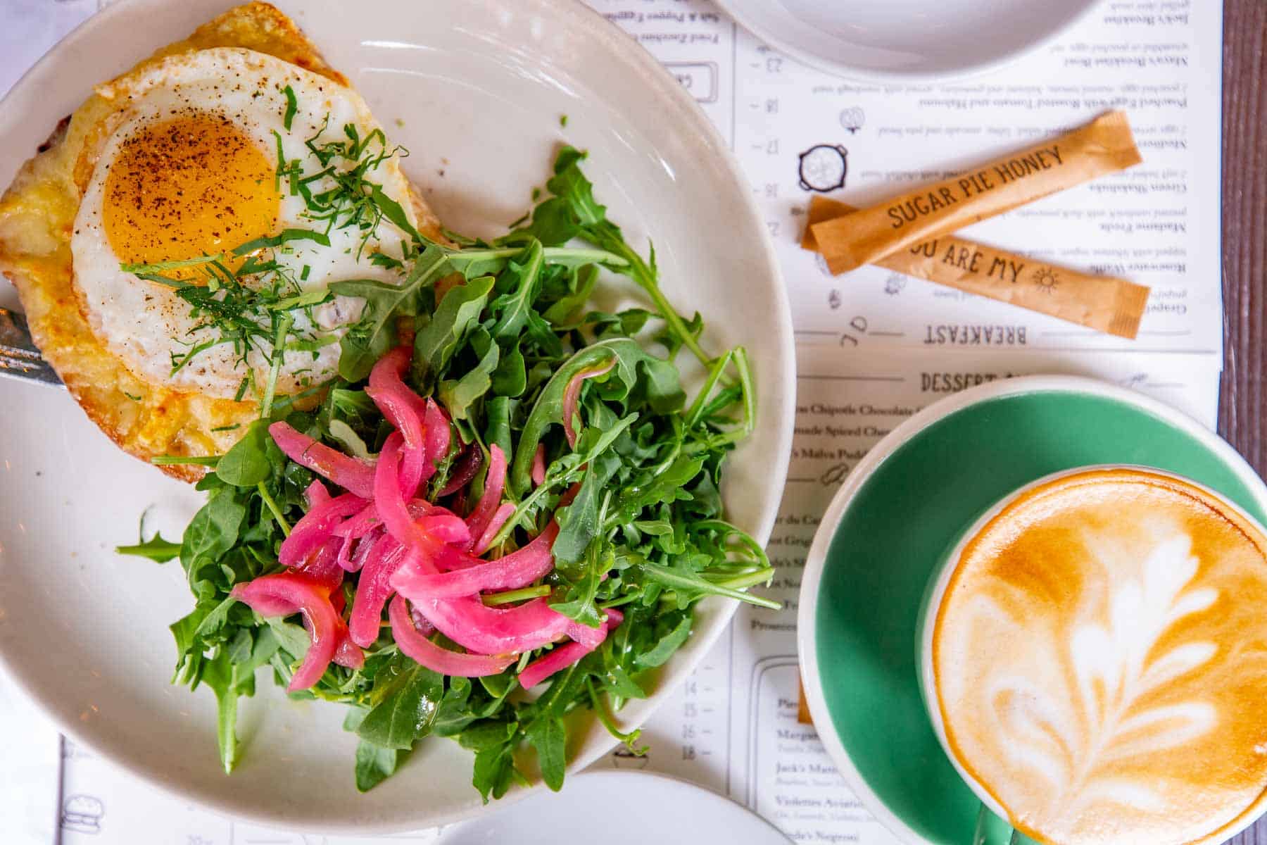 A brunch plate with a fried egg, arugula salad and pickled onions sits beside a latte at Jack's Wife Freda.