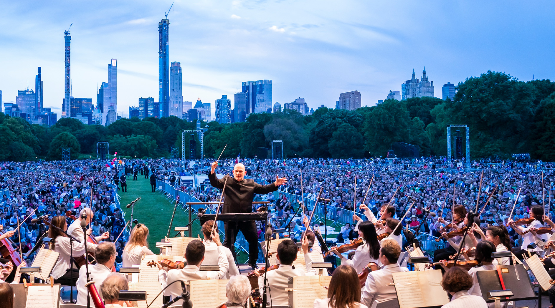 A conductor leads the New York Philharmonic before a massive crowd on Central Park's Great Lawn at dusk.