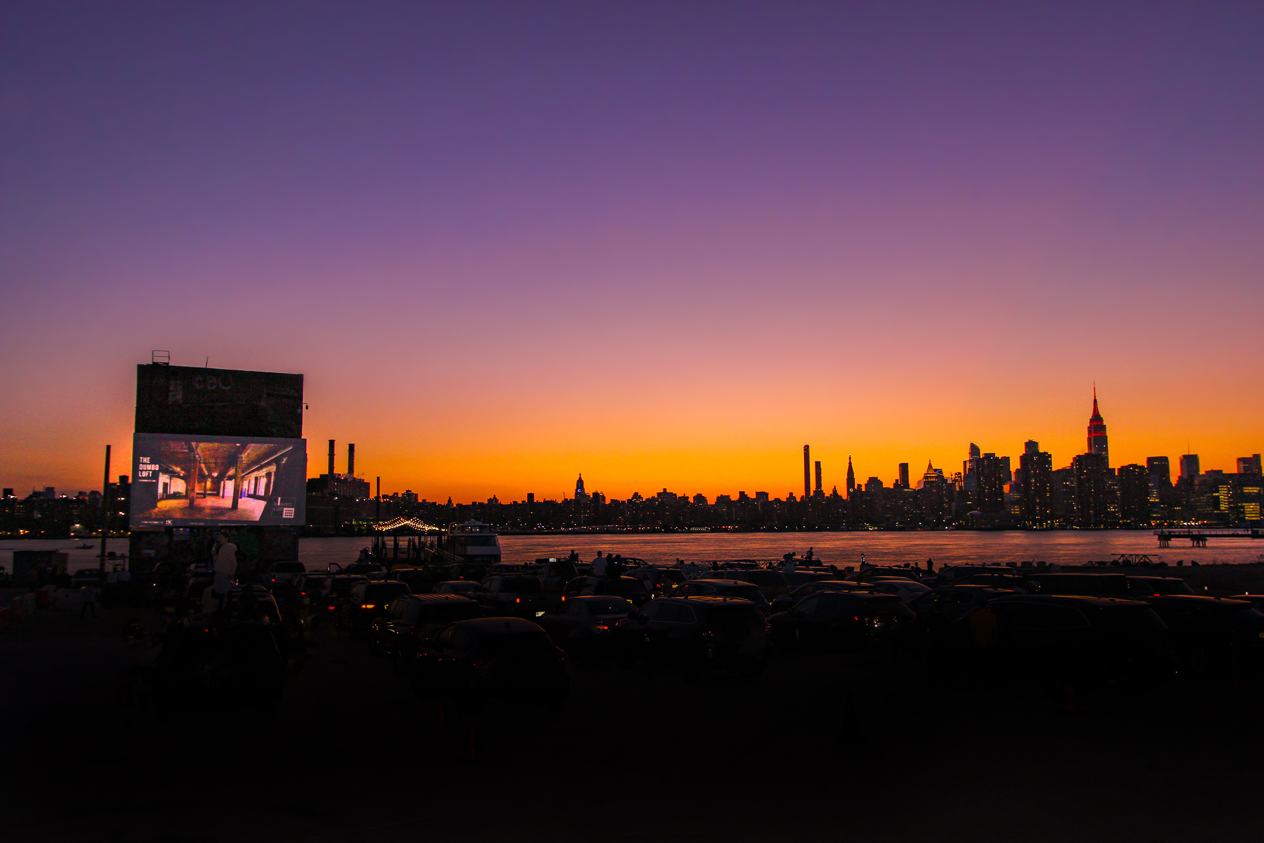 Cars gather at a drive-in movie screening with the Manhattan skyline glowing at dusk across the river.