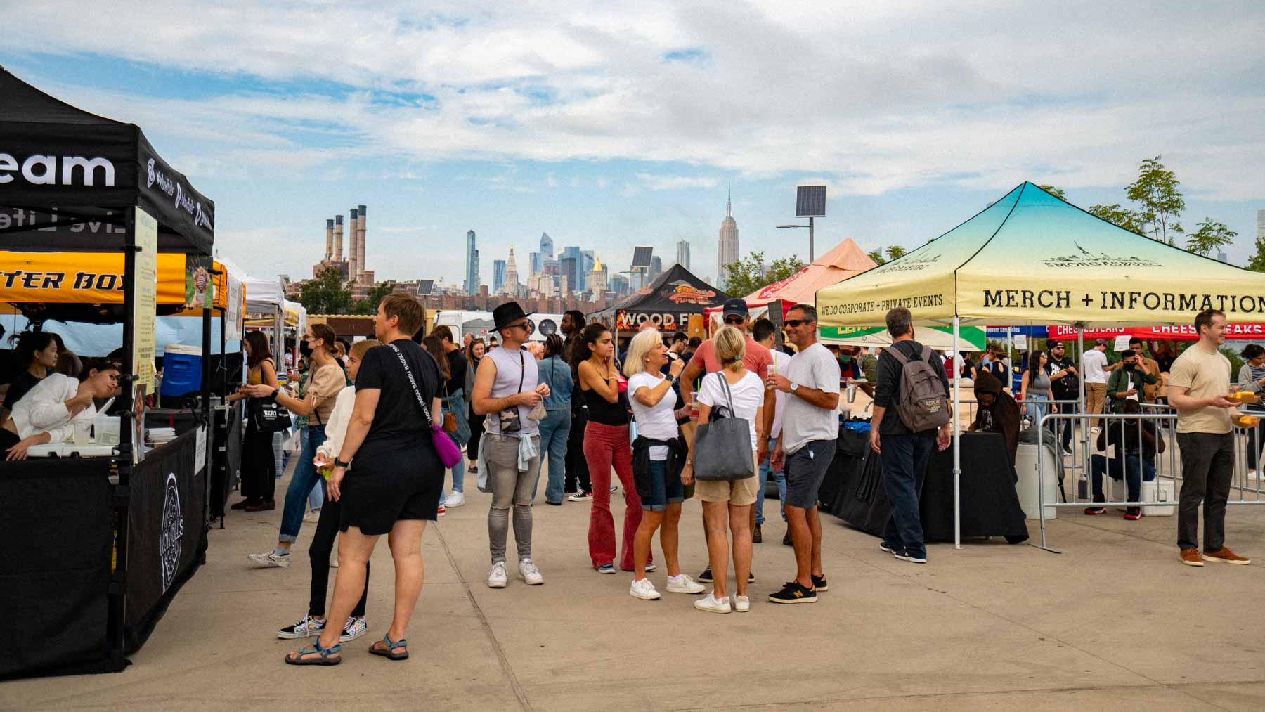 Crowds browse food vendor tents at Smorgasburg with the Manhattan skyline in the background.