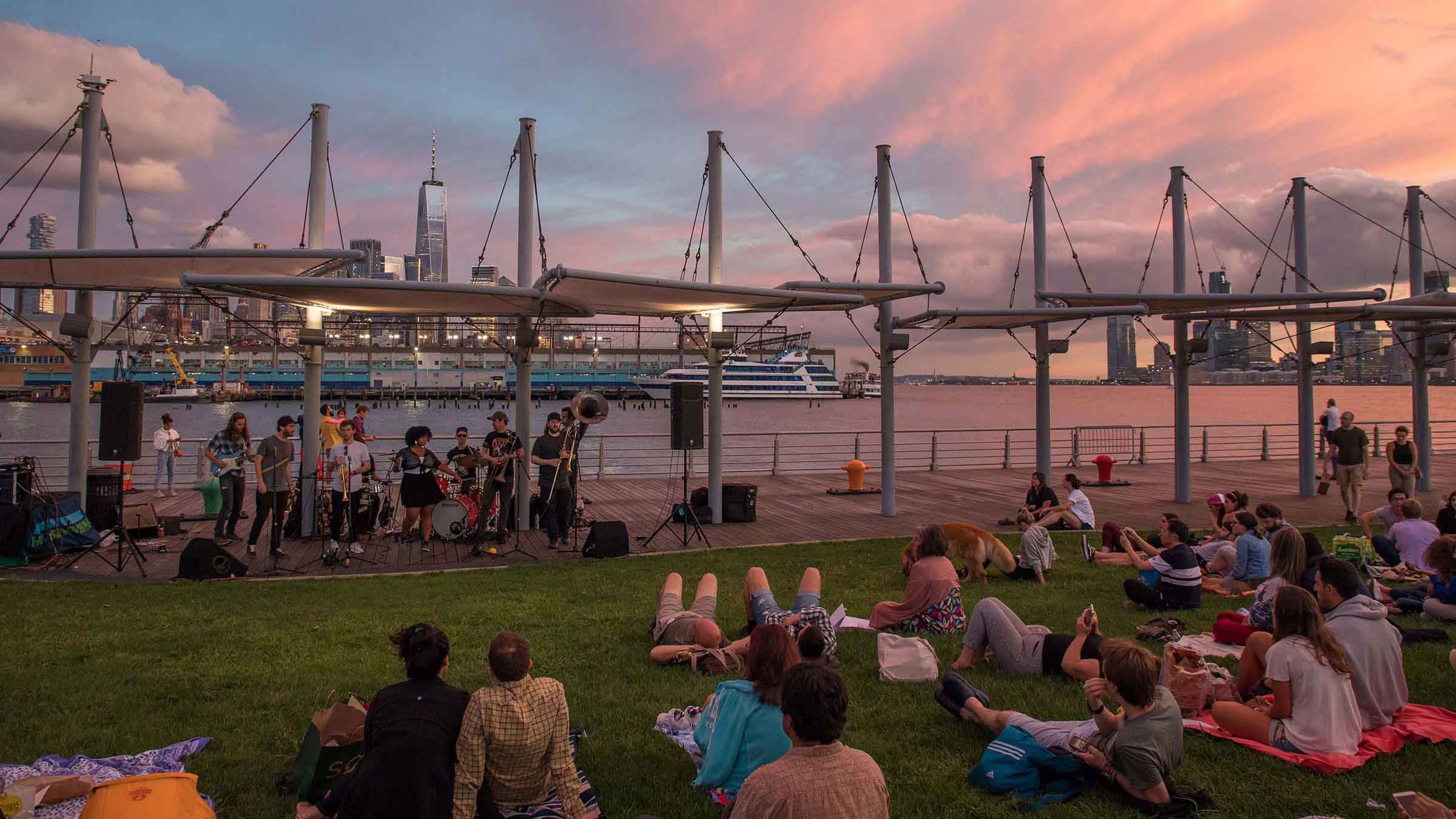 A brass band performs on a pier stage at sunset as people relax on the lawn overlooking the Hudson River and Manhattan skyline.