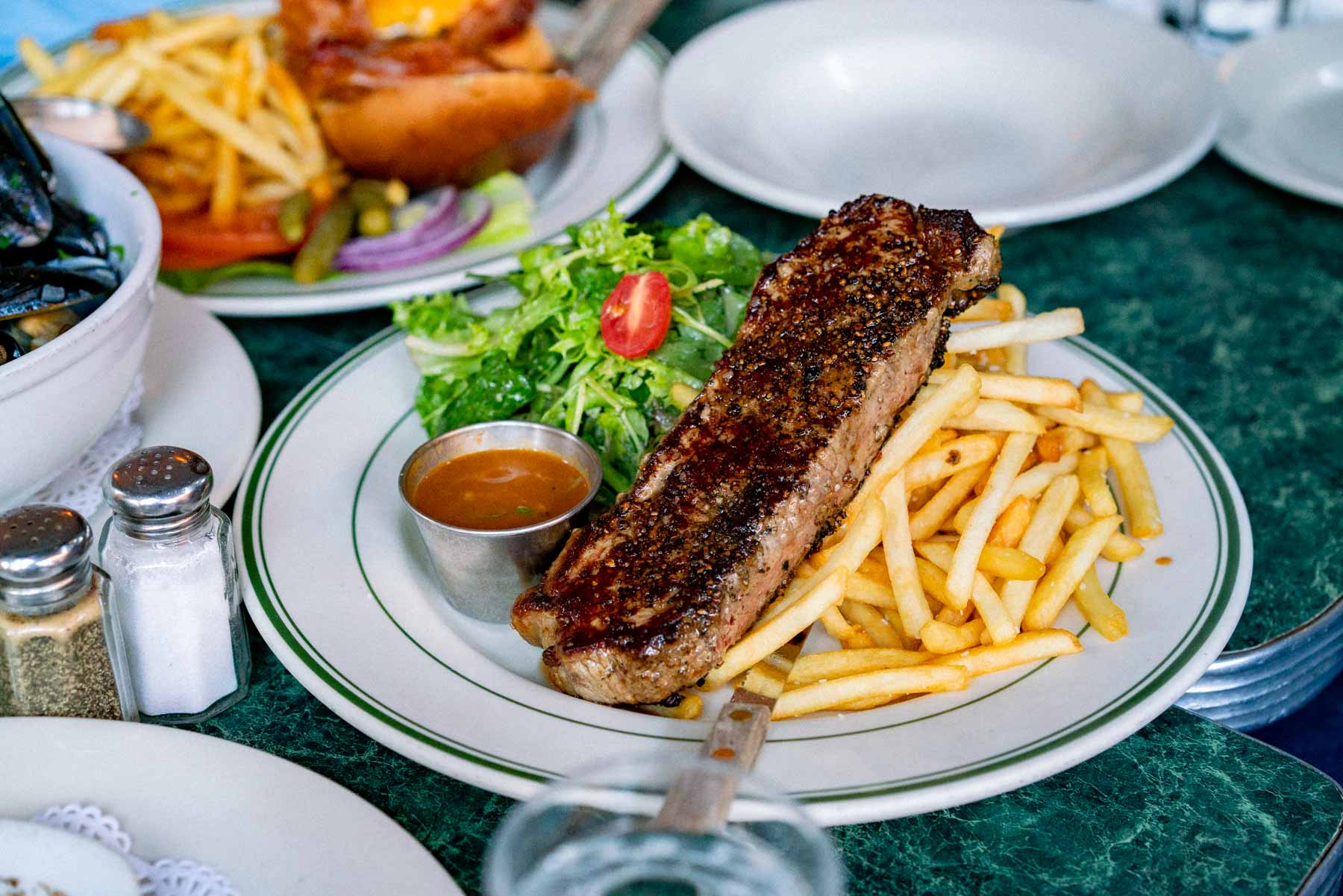 A steak frites plate with salad and peppercorn sauce sits on a table with other dishes.