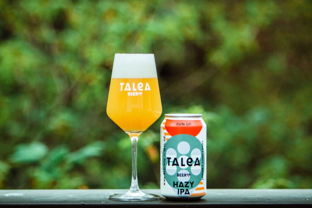 A glass of hazy IPA is poured next to a Talea Beer Co can on a ledge outdoors.