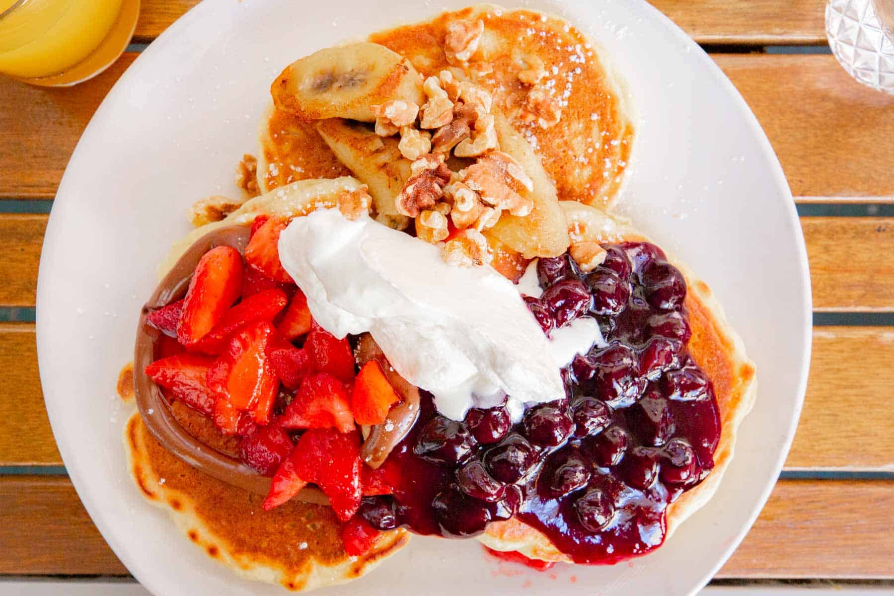 A stack of pancakes topped with strawberries, blueberry compote, bananas, walnuts and whipped cream on a wooden table.