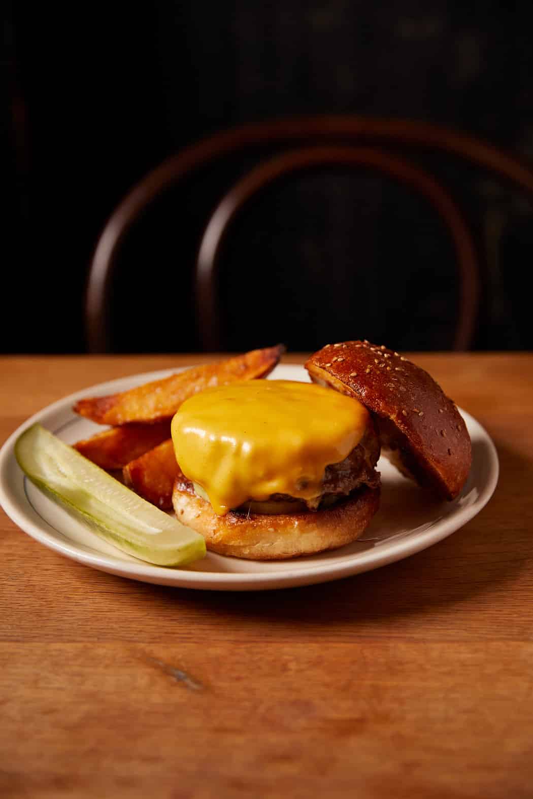 A cheeseburger with melted cheddar sits open-faced on a plate with a pickle spear and potato wedges.