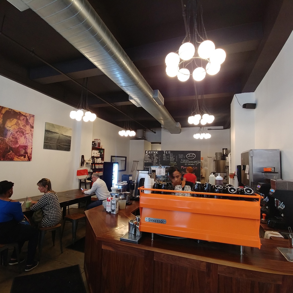 Interior of Cafe Grumpy with an orange espresso machine at the counter and customers seated at a communal table.