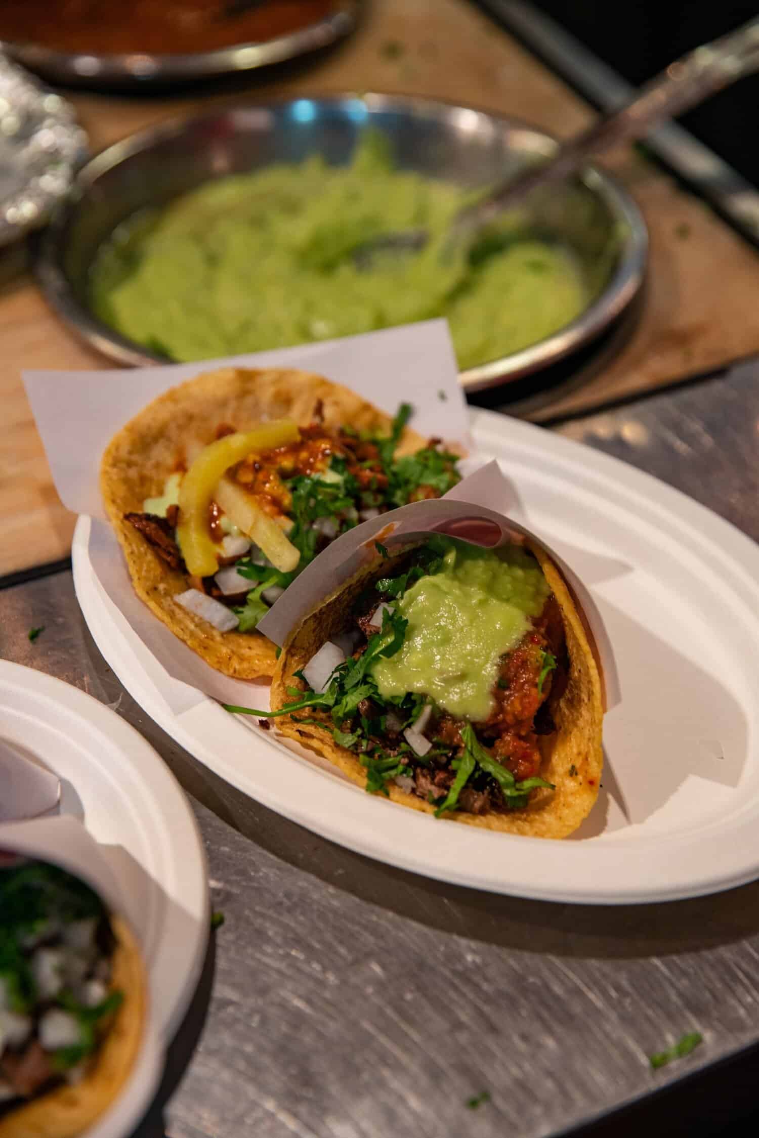 Two carne asada tacos in white paper trays topped with guacamole, salsa, onion and cilantro on a metal counter