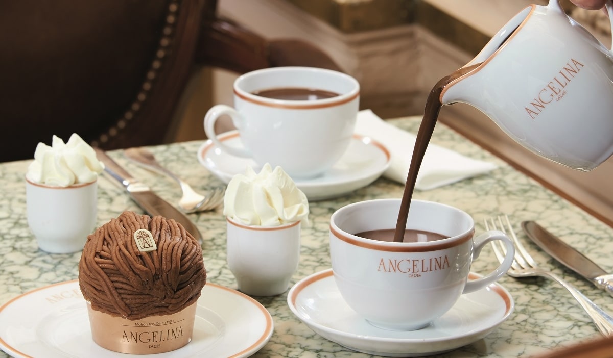 Hot chocolate is poured into a cup beside a Mont Blanc pastry at a marble café table.