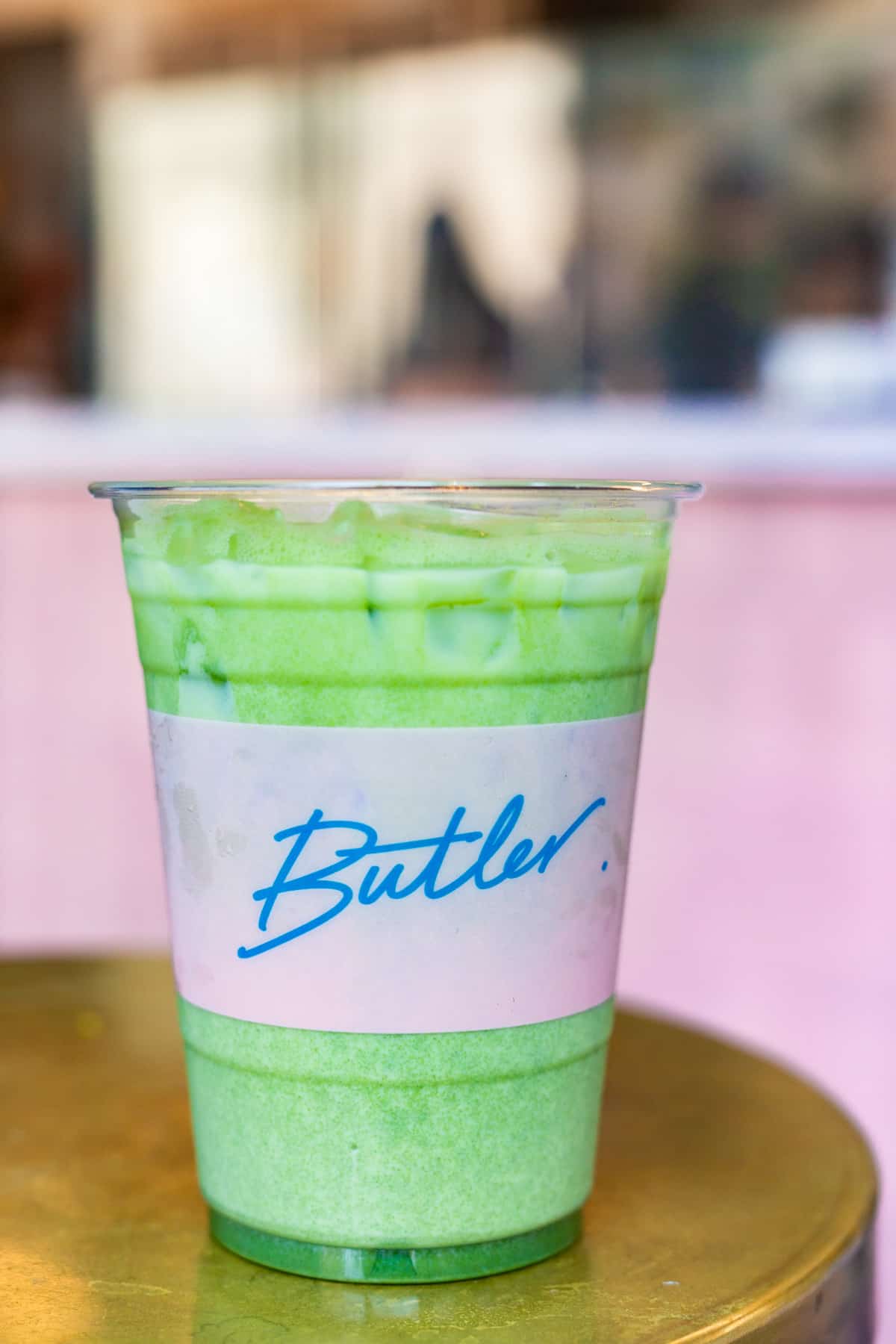 An iced matcha drink in a plastic cup labeled Butler sits on a gold table.