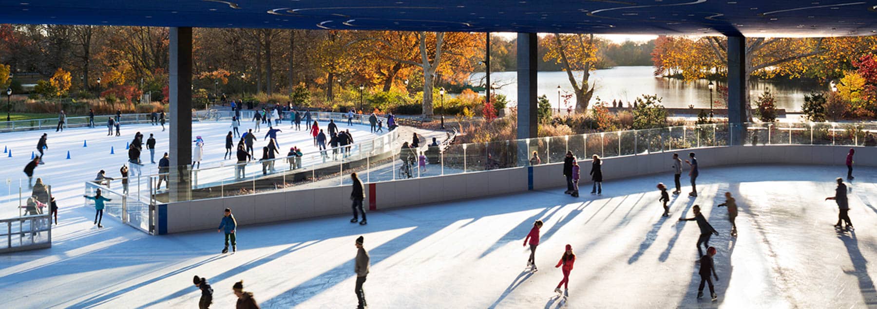 People ice skate at an outdoor rink beside a lake in a park with fall foliage