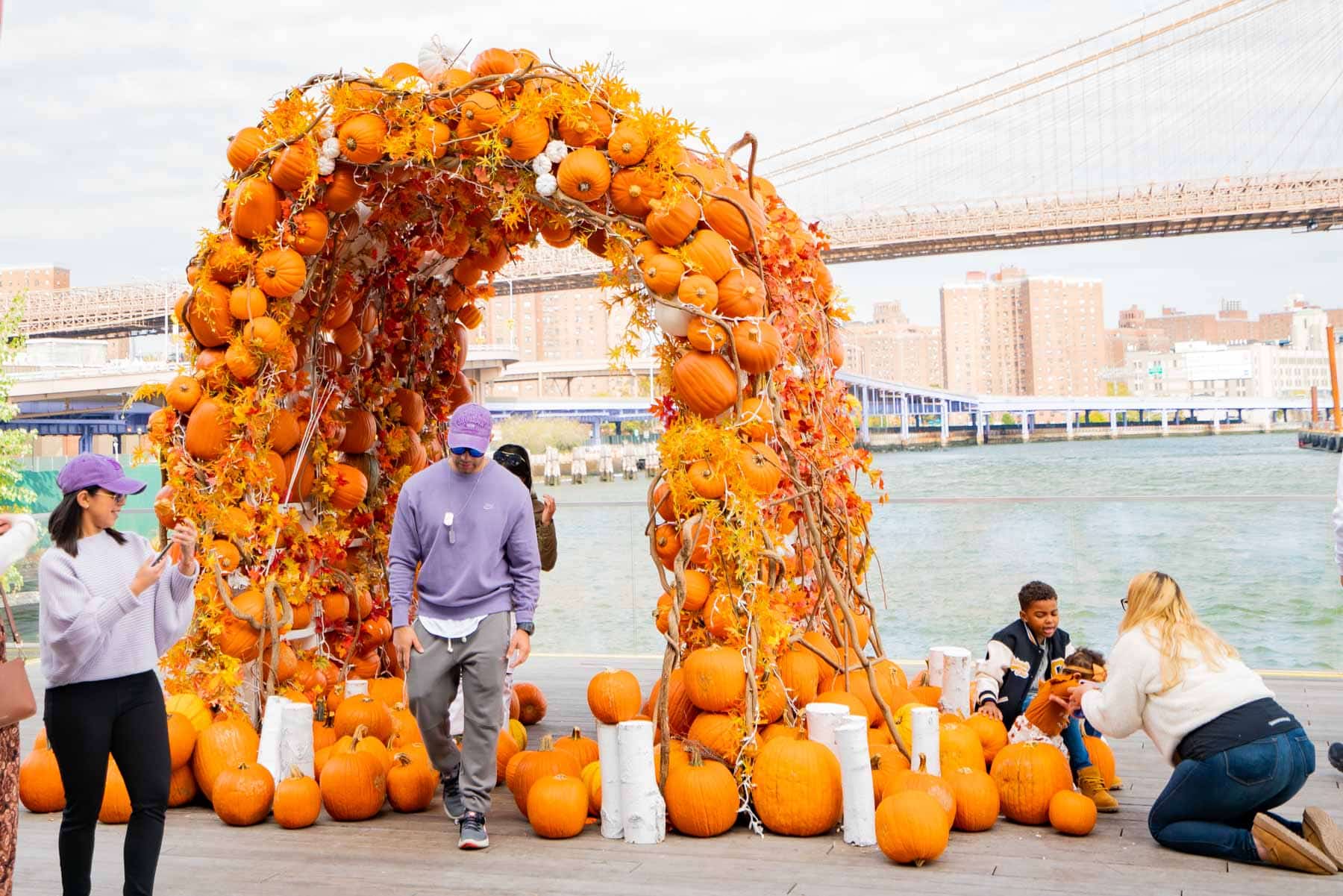 People gather around a large pumpkin arch installation near the East River with the Brooklyn Bridge in the background.