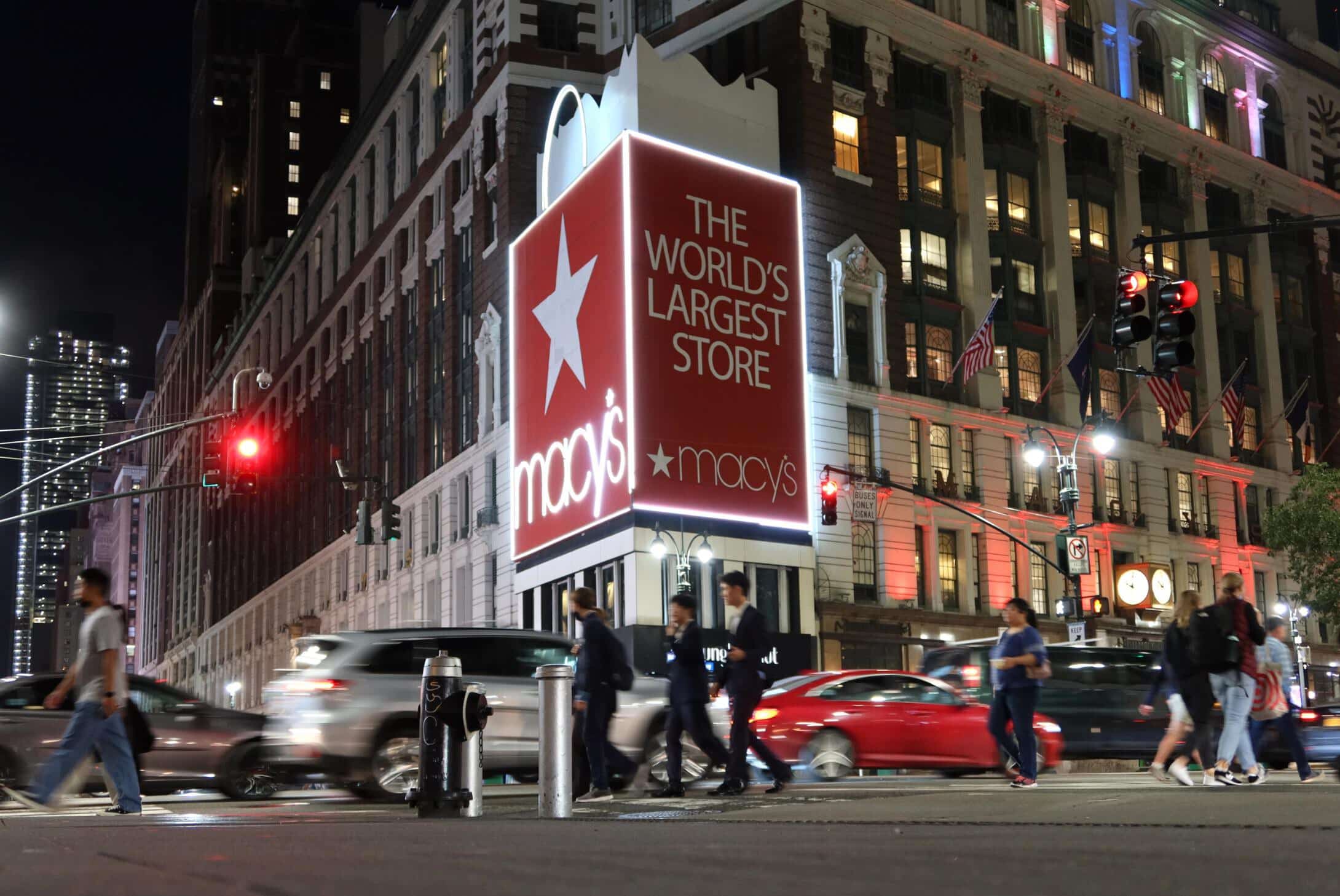 Pedestrians cross the street at night beneath the illuminated Macy's sign at Herald Square.