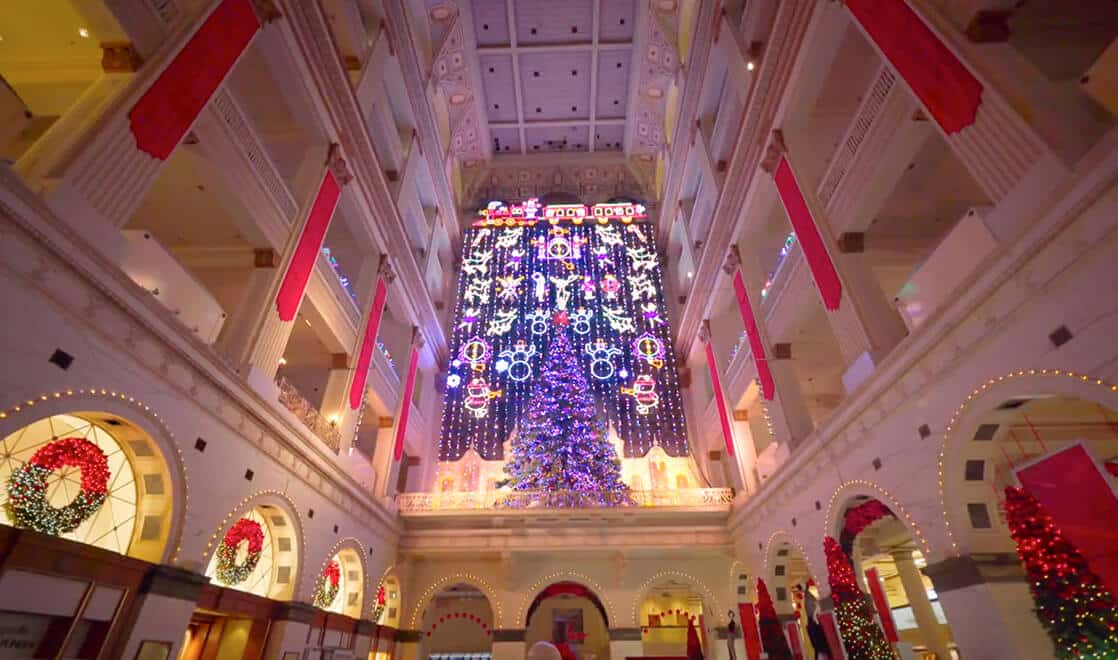 A grand atrium inside a department store is decorated with Christmas trees, wreaths, and light displays.