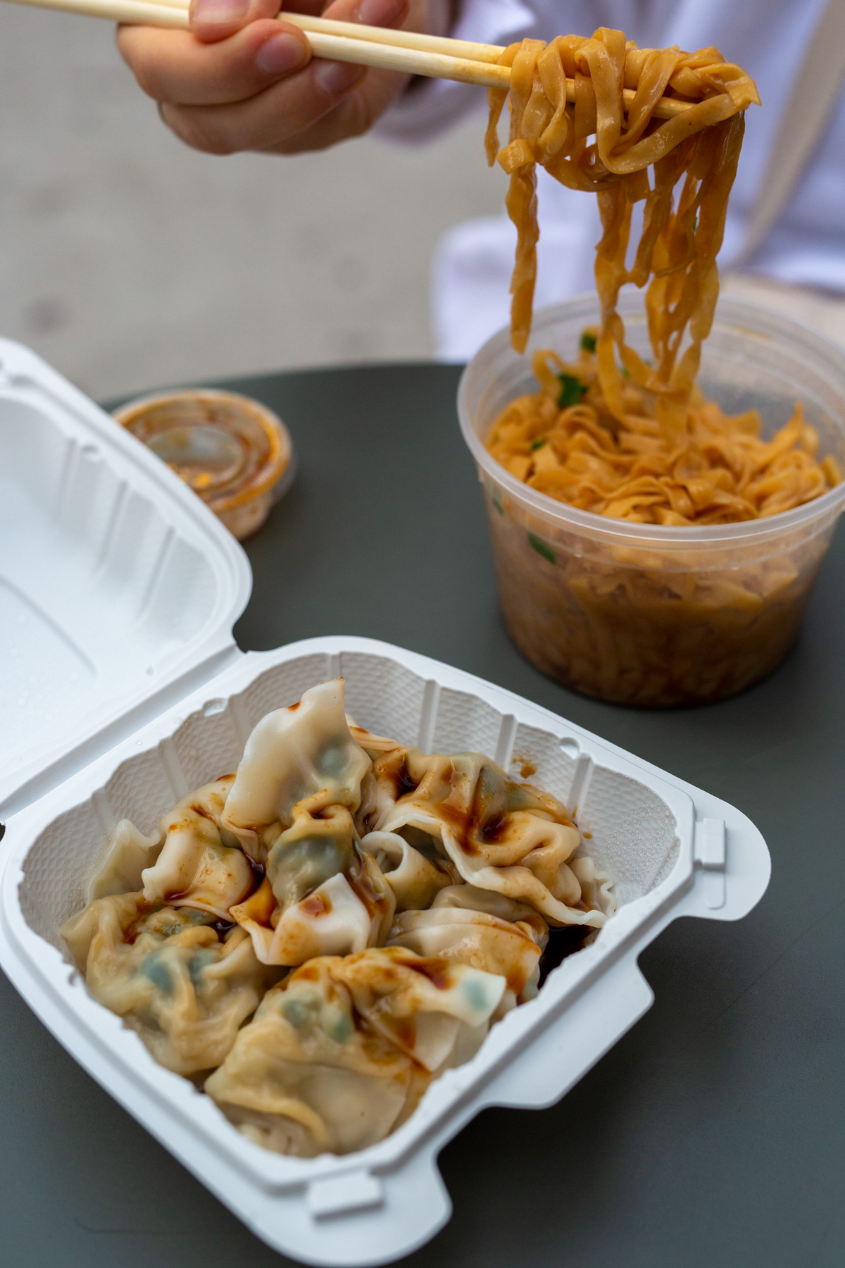 Pork and vegetable dumplings in a takeout box with chili-oil hand-pulled noodles lifted on chopsticks from a plastic cup