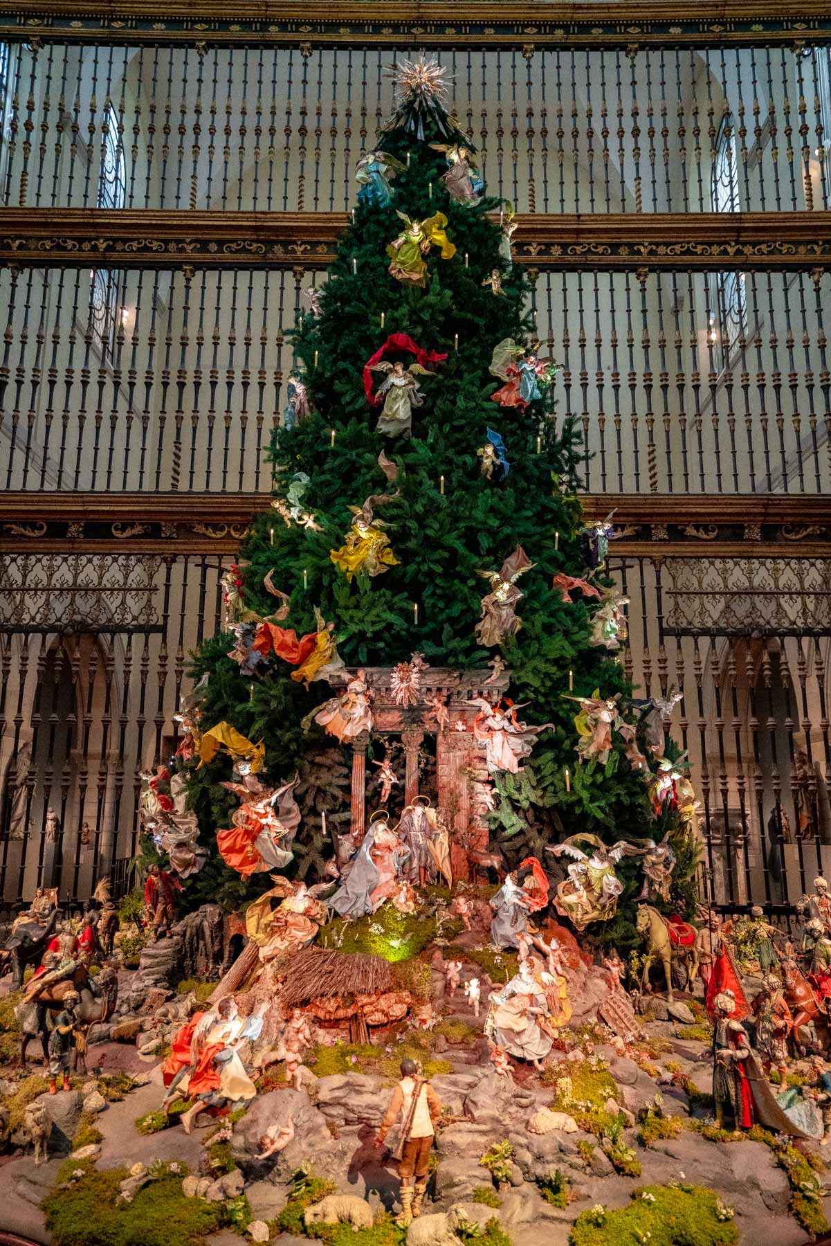 A large Christmas tree decorated with angels stands above a Neapolitan nativity scene inside a museum gallery.