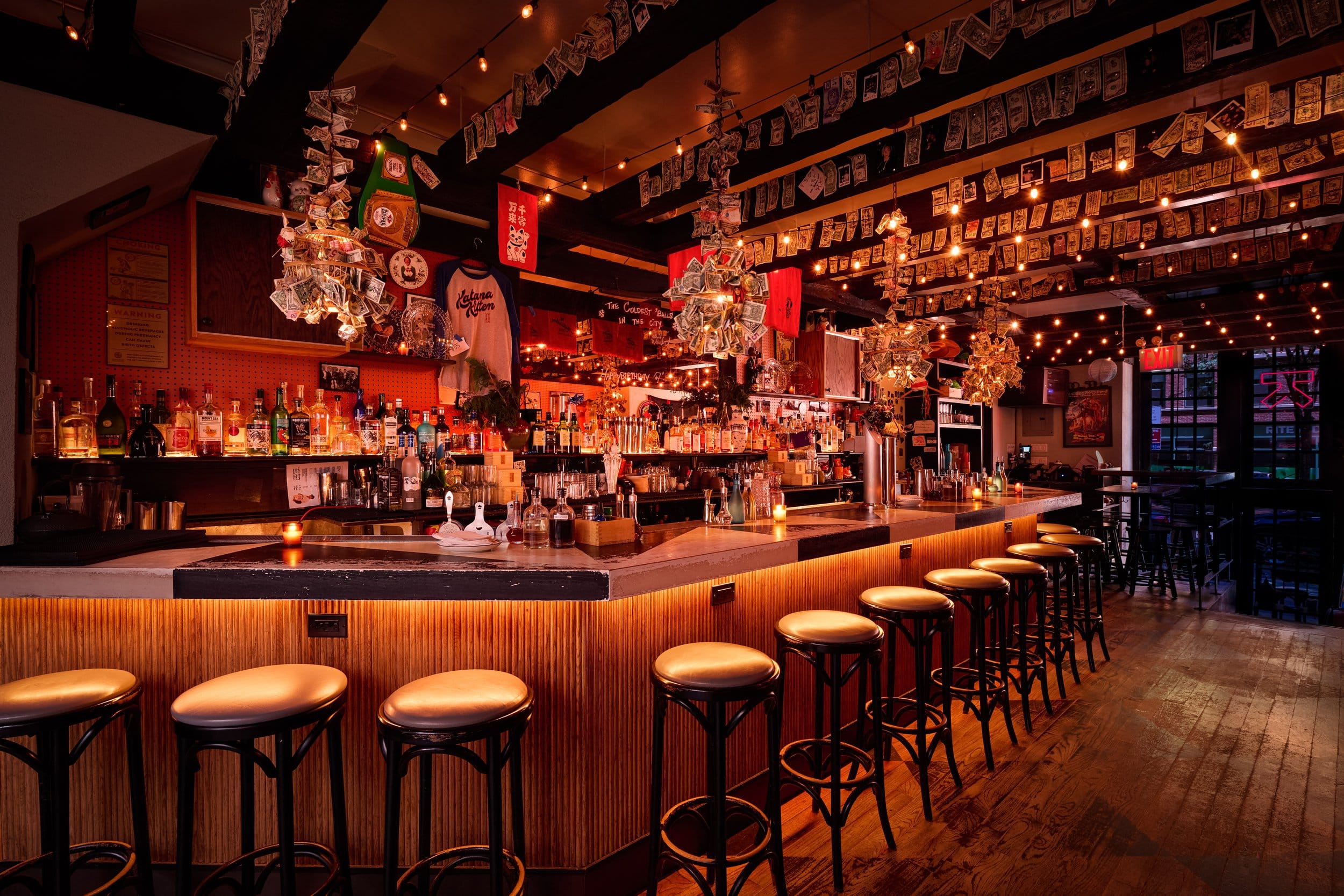 An empty bar interior with wooden stools lines a long counter beneath hanging decorations and string lights.