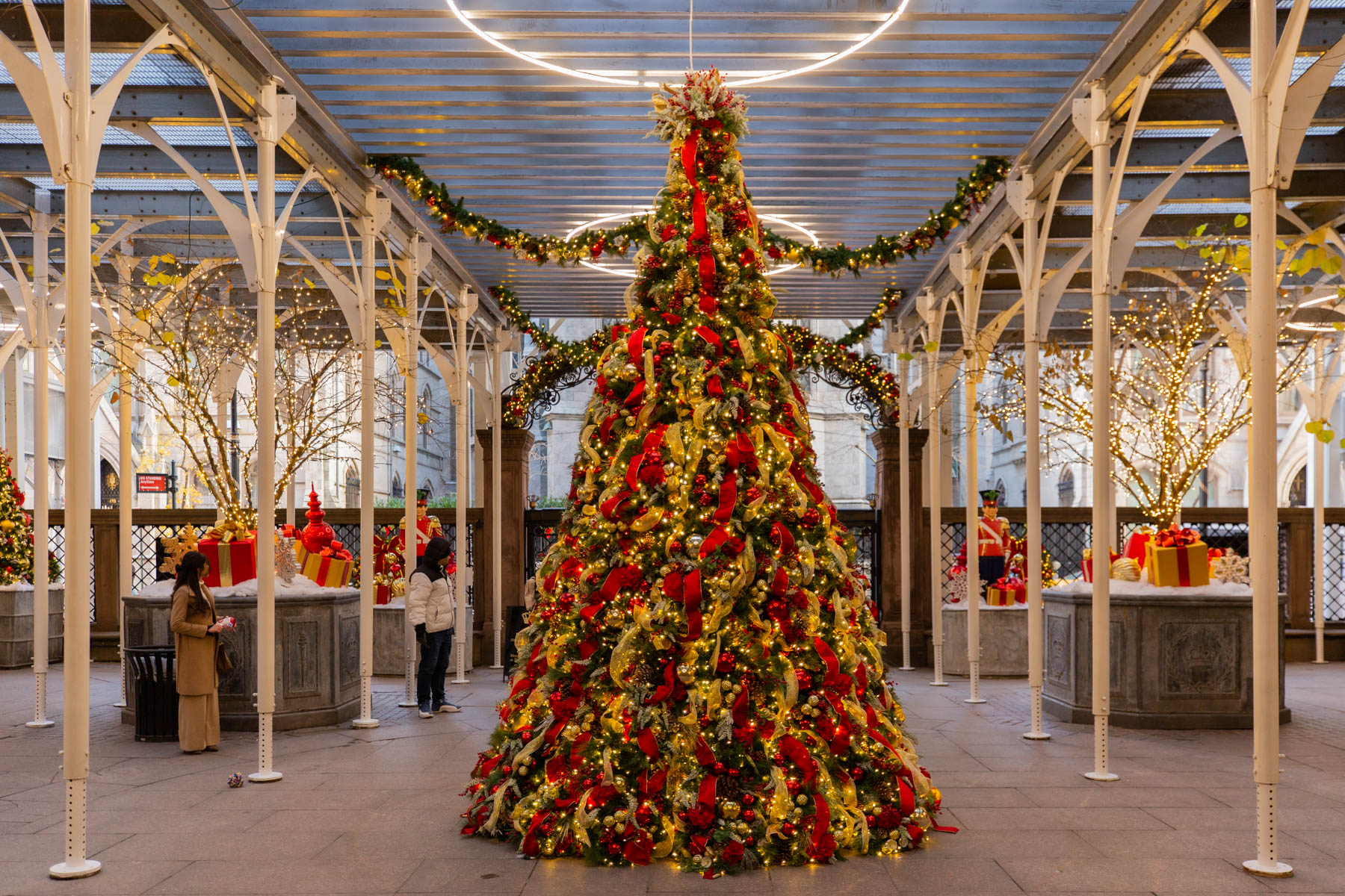 A large red and gold Christmas tree stands beneath a glass and iron courtyard structure decorated for the holidays.