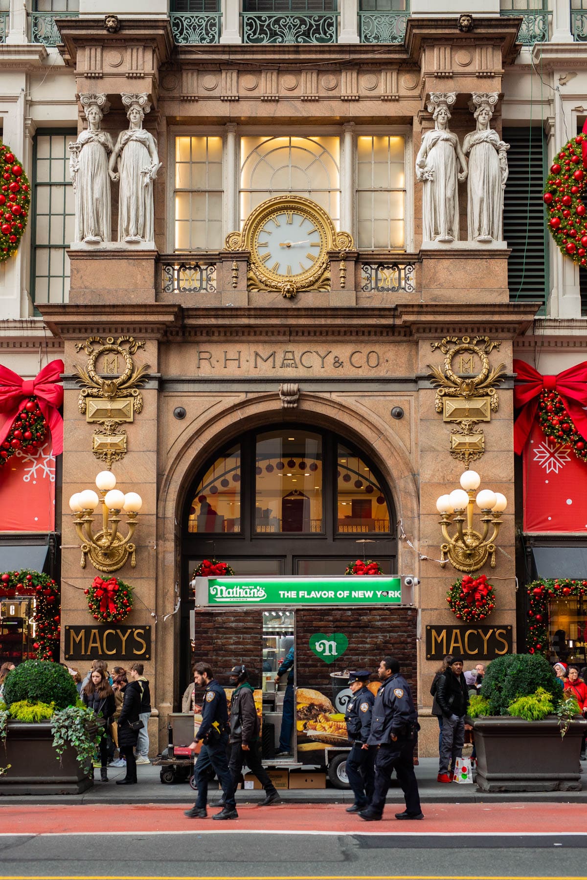 The R.H. Macy & Co. building entrance is decorated with Christmas wreaths and bows above a Nathan's food cart on the sidewalk.