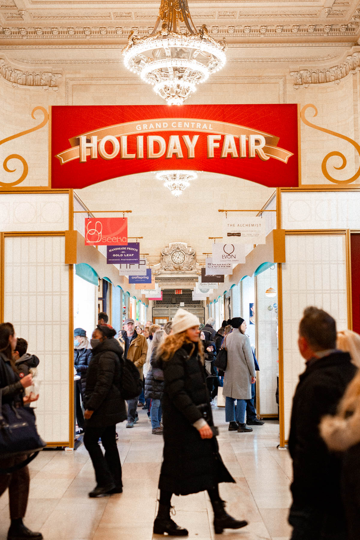 Shoppers walk through the Grand Central Holiday Fair beneath a chandelier and ornate archway.