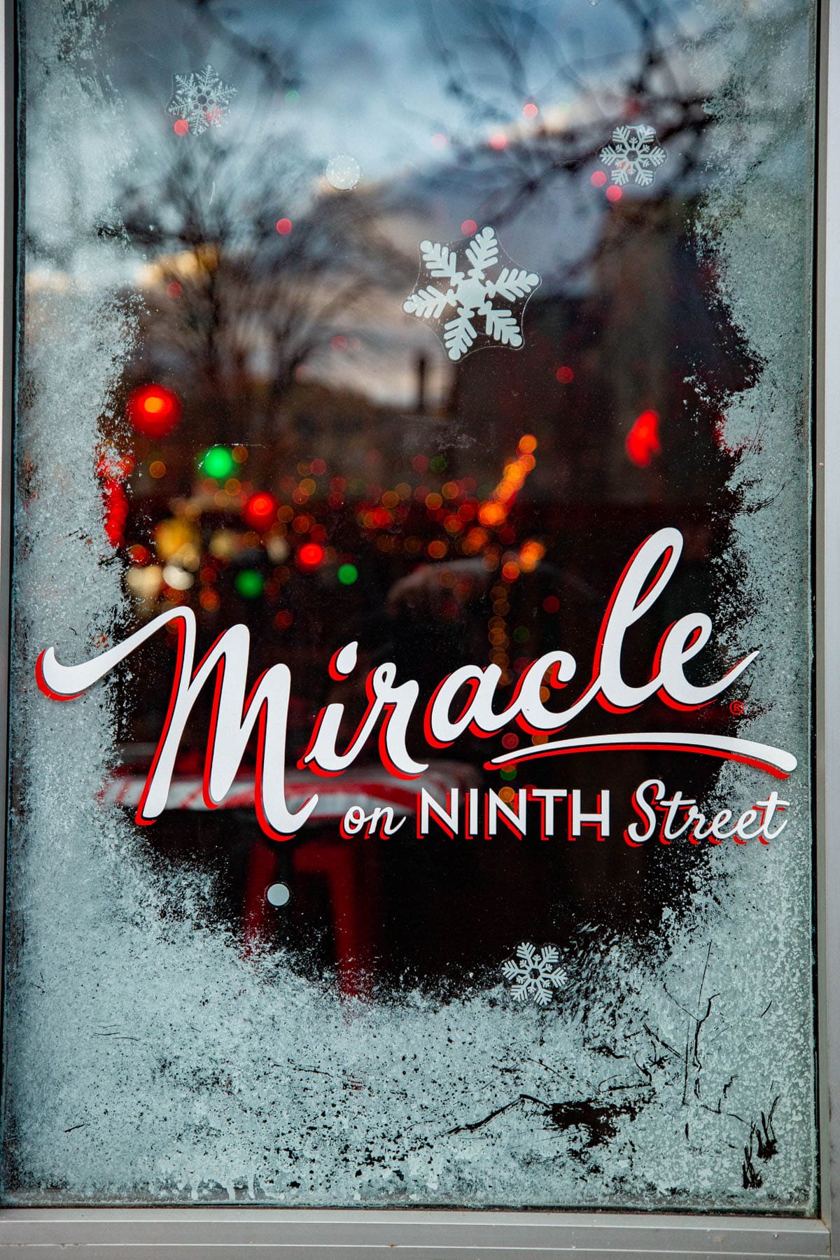 Miracle on 9th Street, a frosted window is decorated with snowflake stickers and a Miracle on Ninth Street sign, with holiday lights glowing inside.