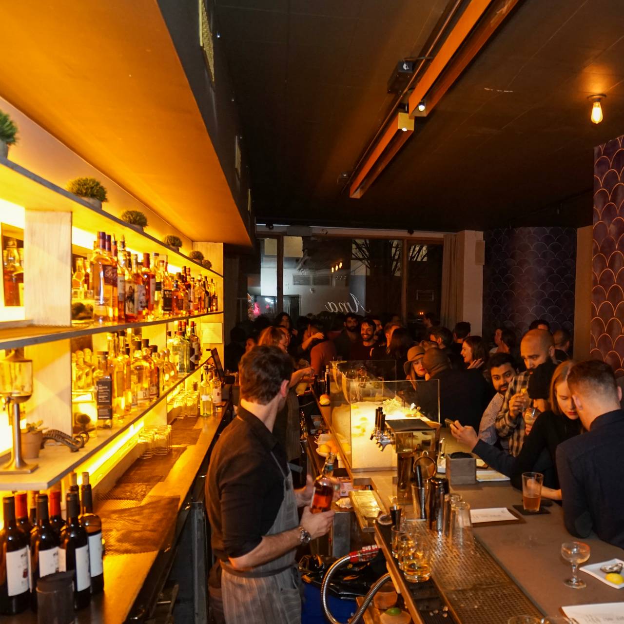 A crowded bar with a bartender pouring drinks and shelves of liquor bottles lit from below.