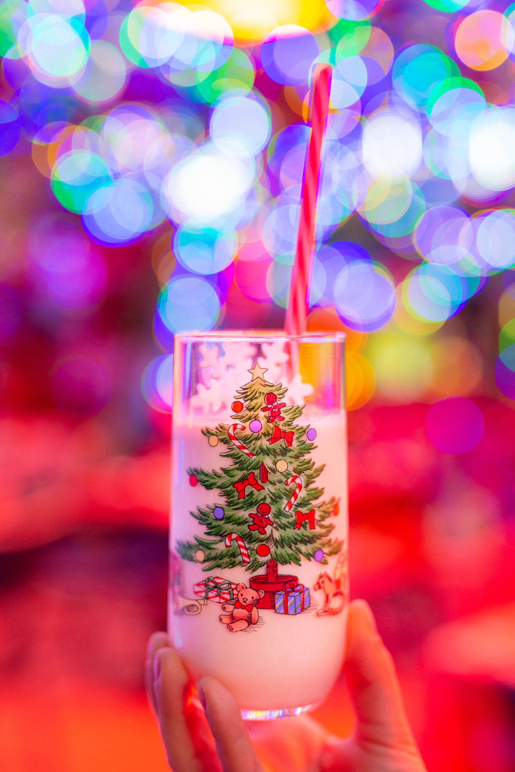 A hand holds a festive milky cocktail in a Christmas-tree printed glass against blurred colorful holiday lights.
