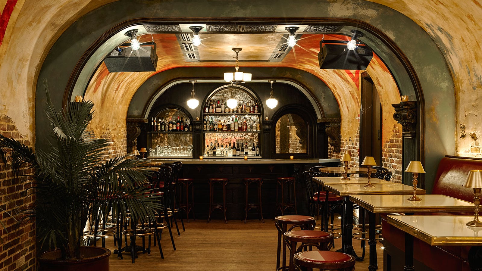 An arched brick basement bar with bottle-lined shelves and marble tables.