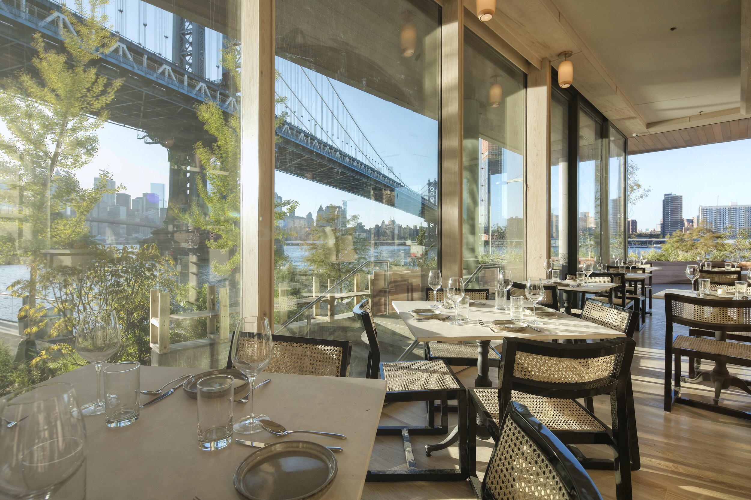 A restaurant dining room with set tables overlooks the Manhattan Bridge and river through floor-to-ceiling windows.
