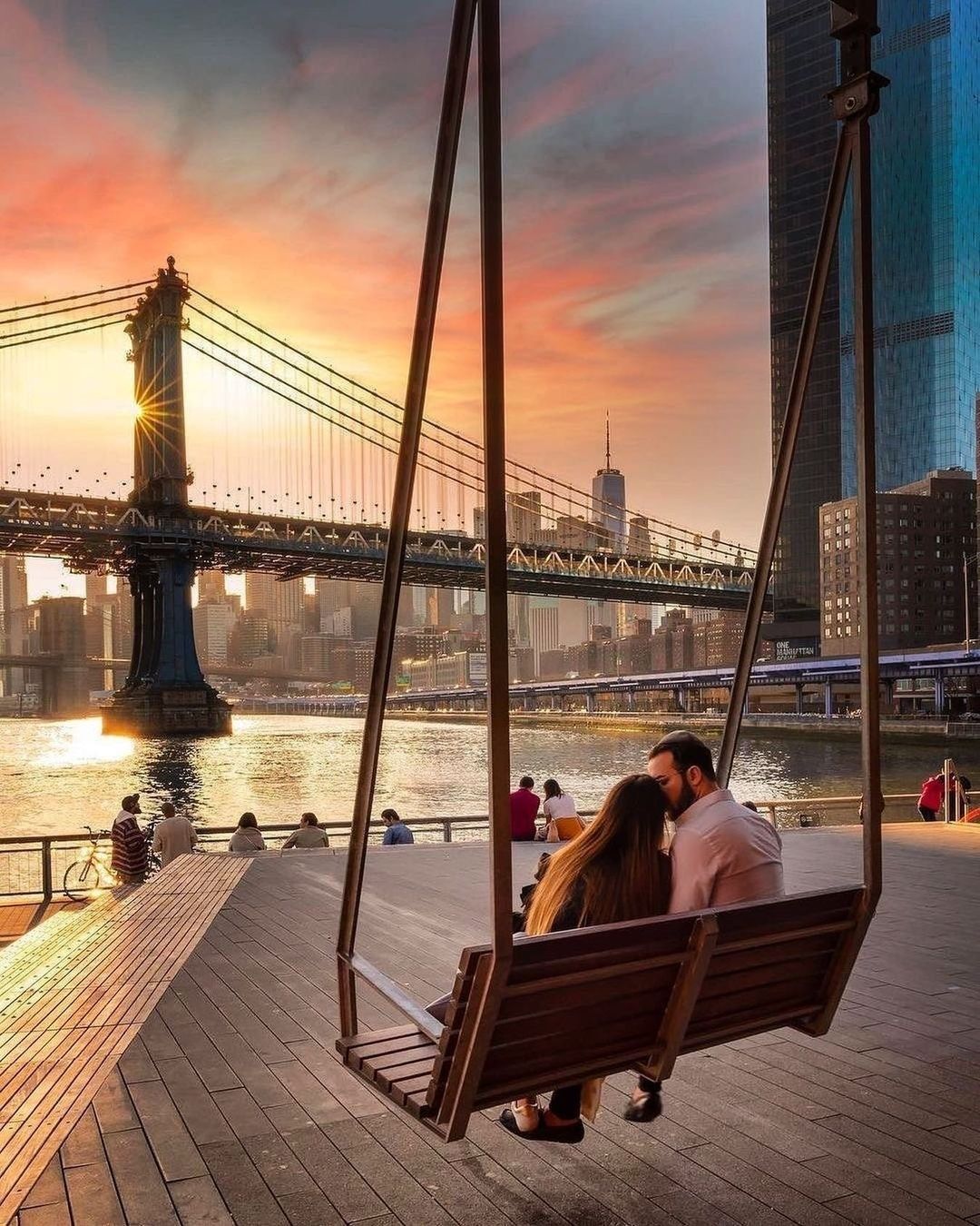 A couple sits on a swing overlooking the Manhattan Bridge and skyline at sunset.