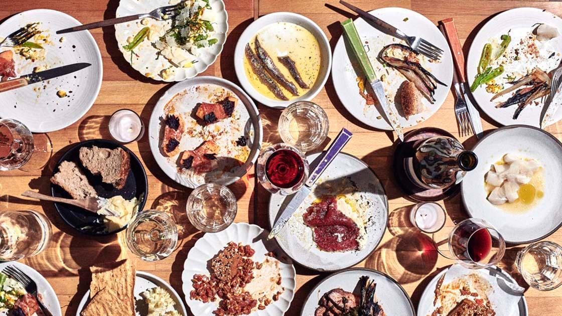 An overhead view of a wooden table covered with many shared plates of half-eaten food and glasses of wine.