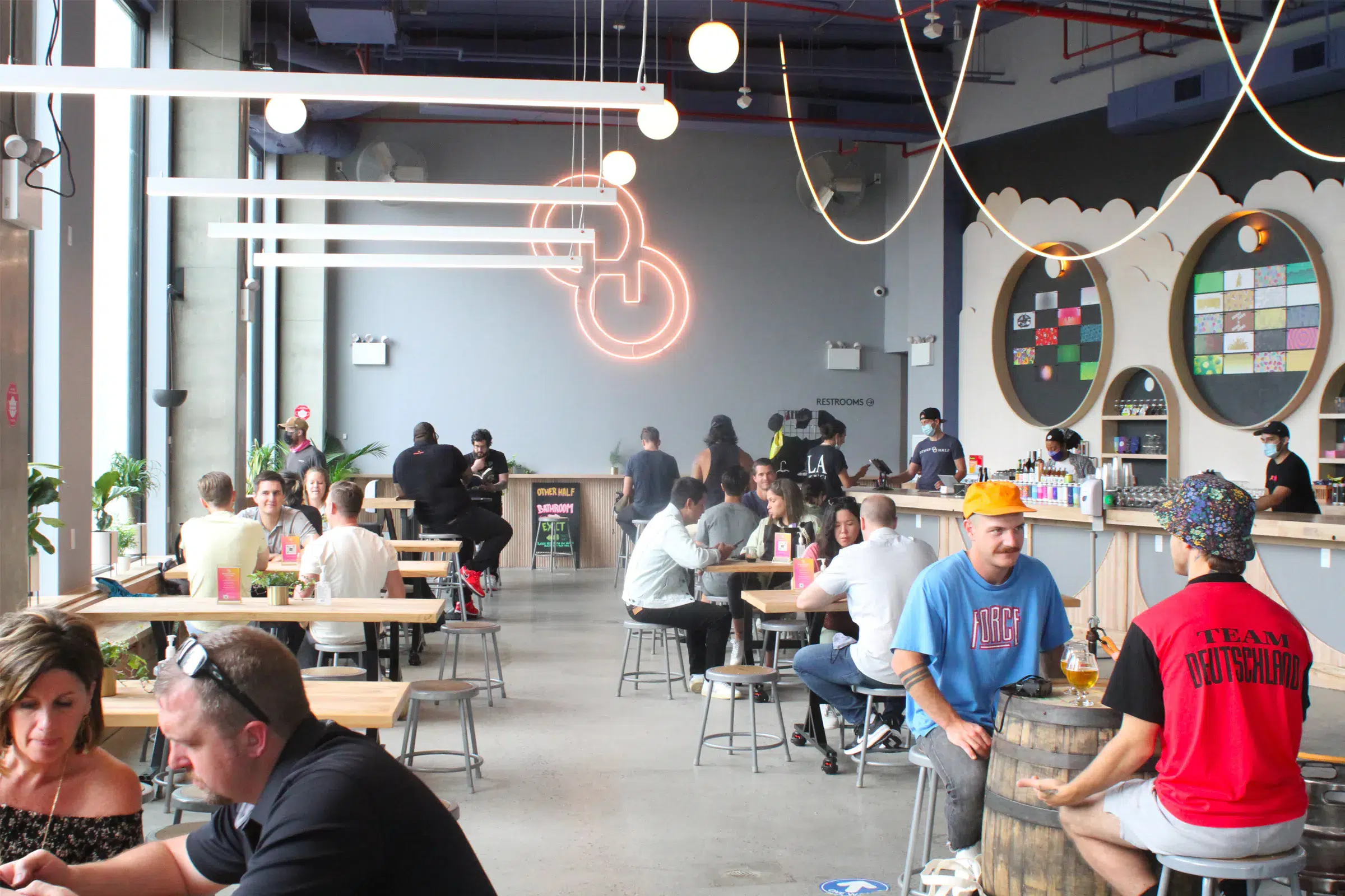People sit at tables and a bar inside a modern brewery taproom with neon signage.