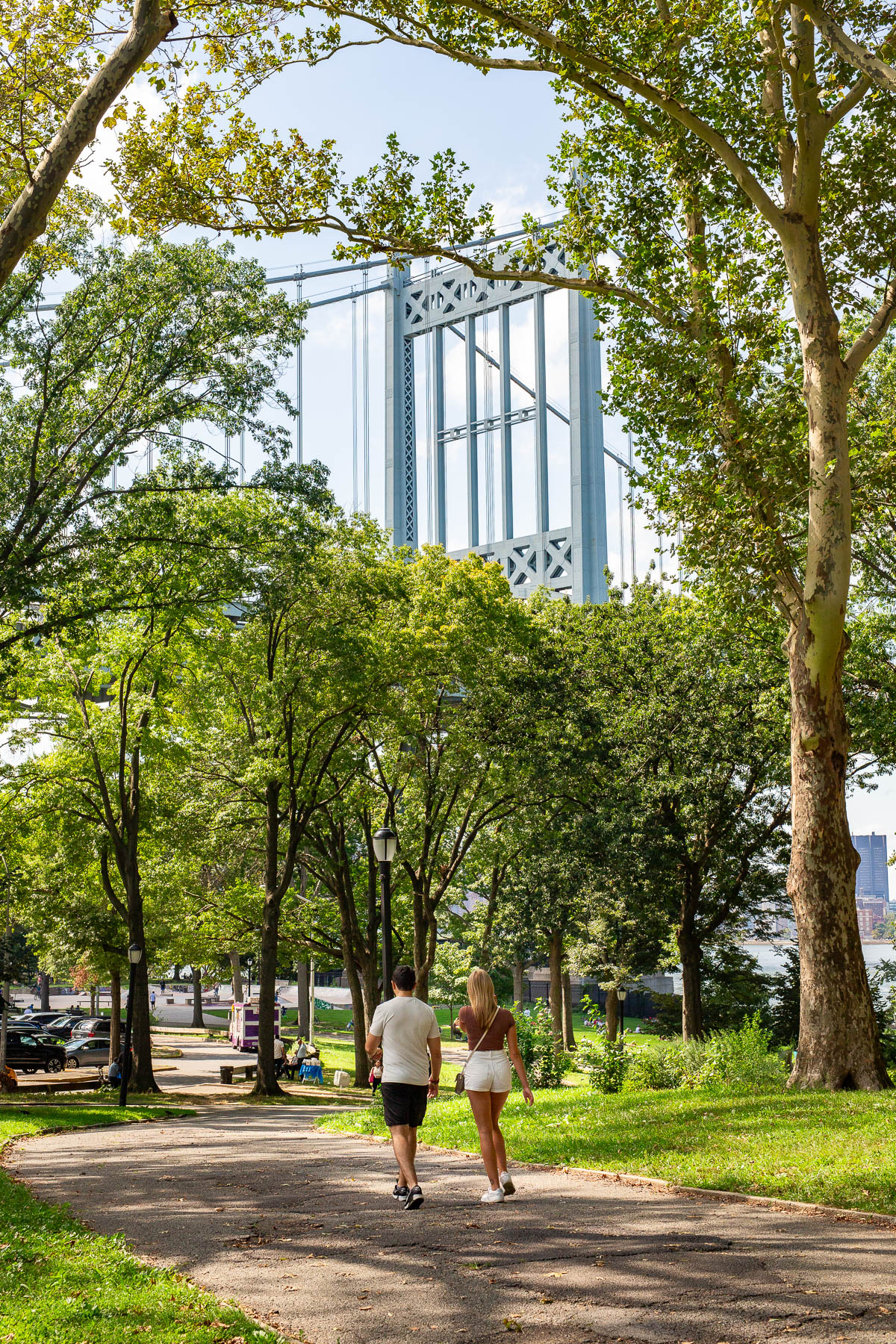 Two people walk along a tree-lined path in Astoria Park with the Hell Gate Bridge visible overhead.