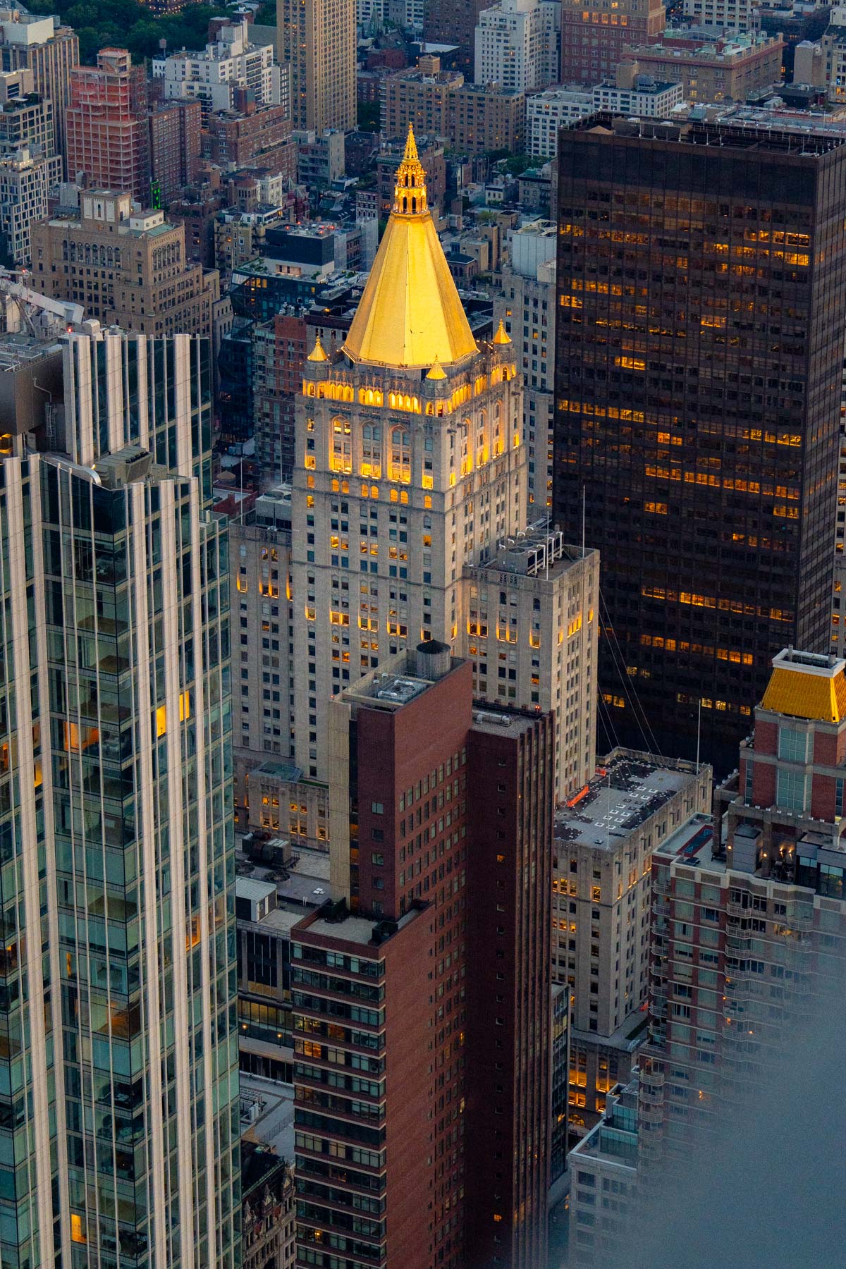 An aerial view of Manhattan skyscrapers at dusk with a gold-topped tower illuminated in the center.